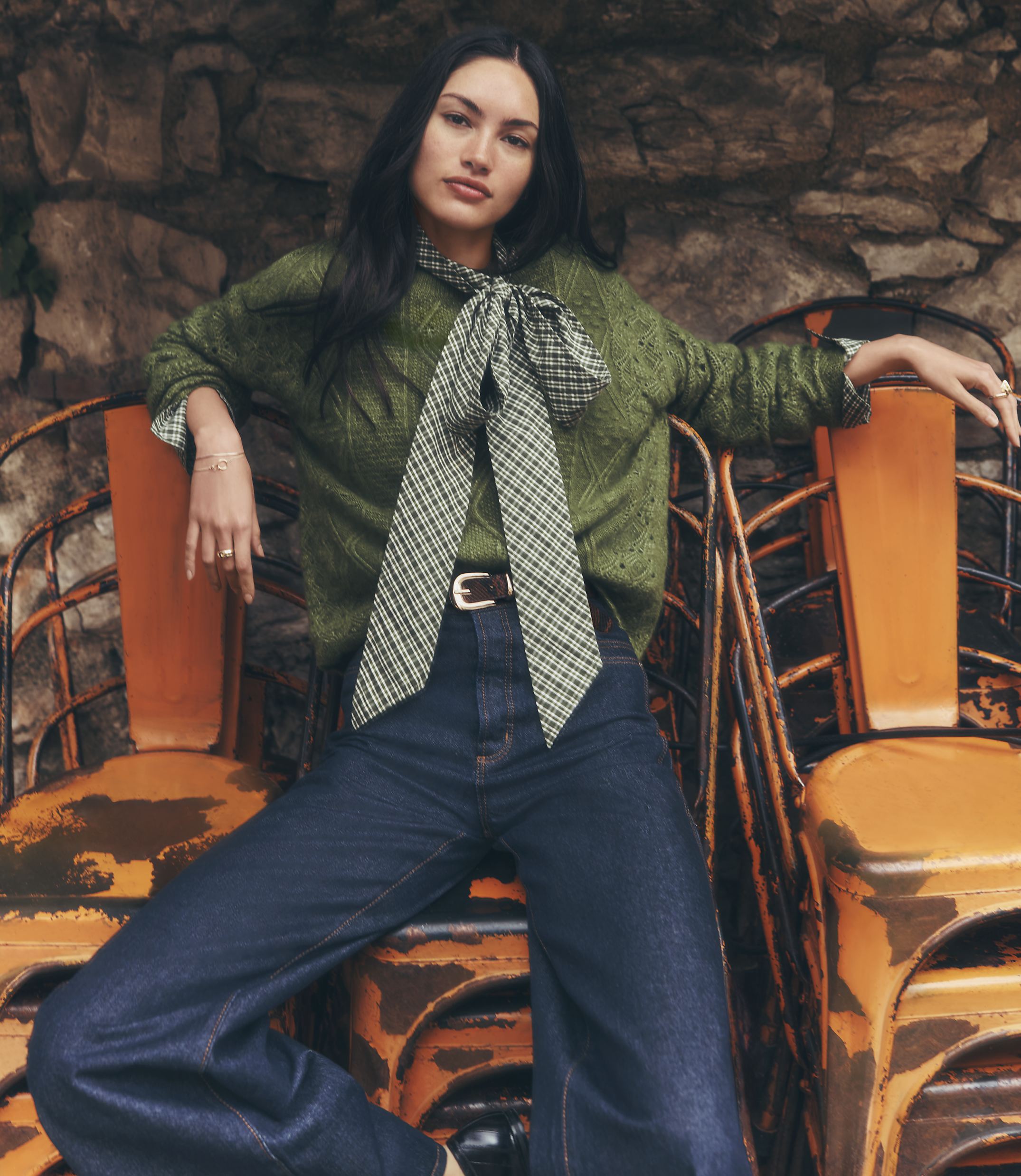 A woman in a green sweater and dark jeans poses in front of a rustic stone wall and stacked orange chairs.