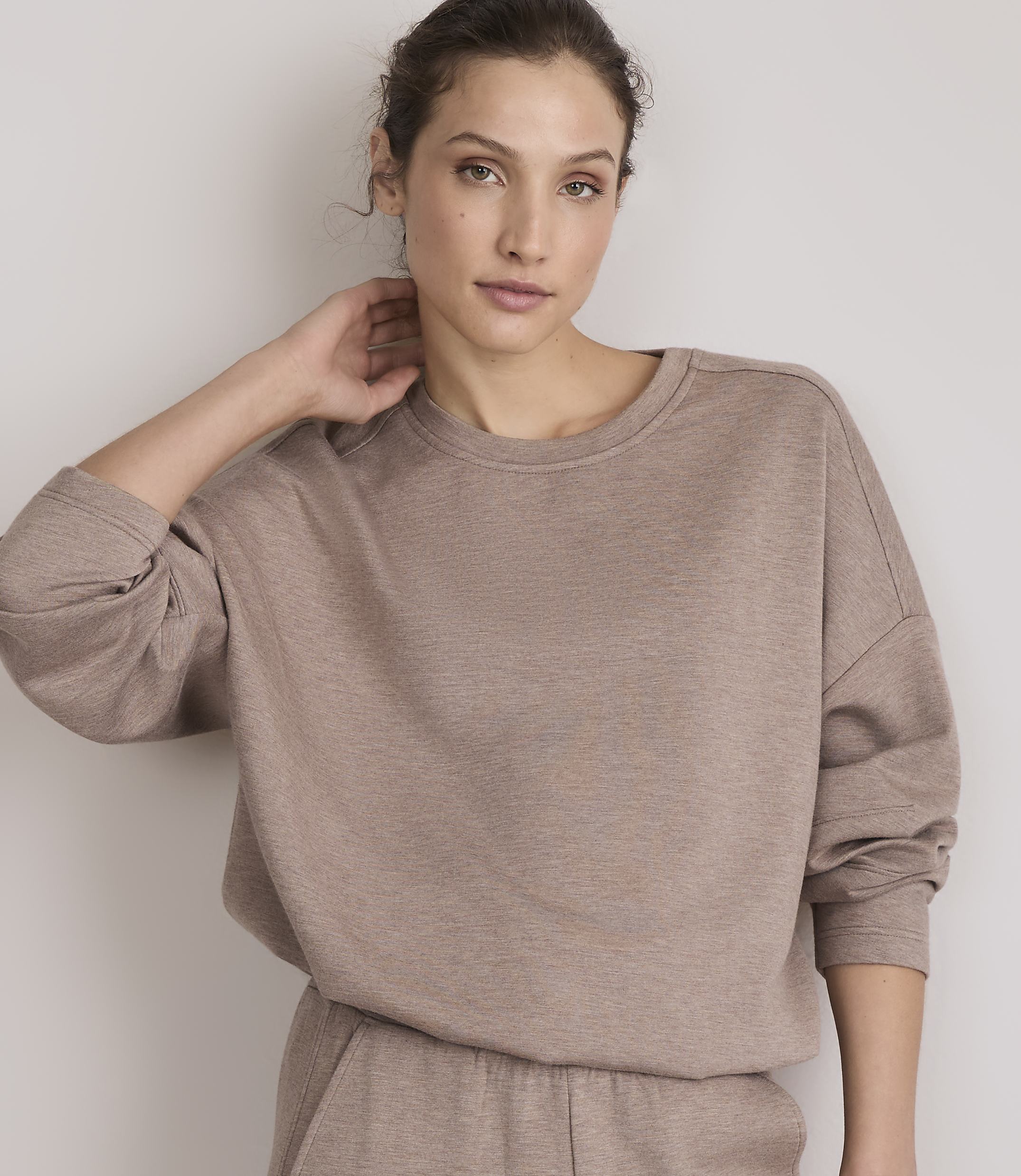 A woman in a taupe sweatshirt and matching pants poses against a plain, light-colored wall.