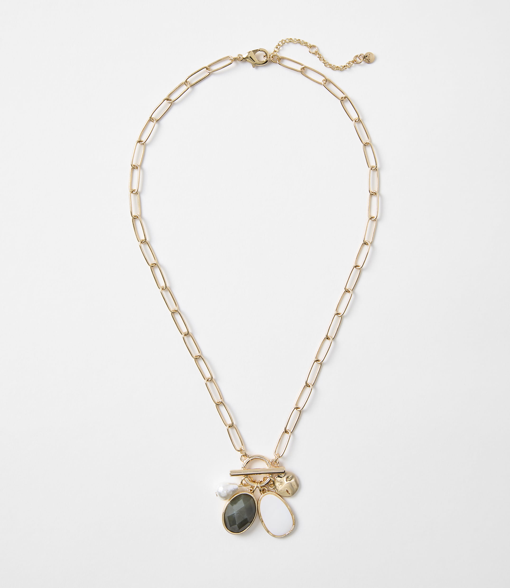A gold paperclip chain necklace with a toggle clasp hangs against a plain white background, featuring three charms: a pearl, a dark oval stone, and a white teardrop-shaped stone.
