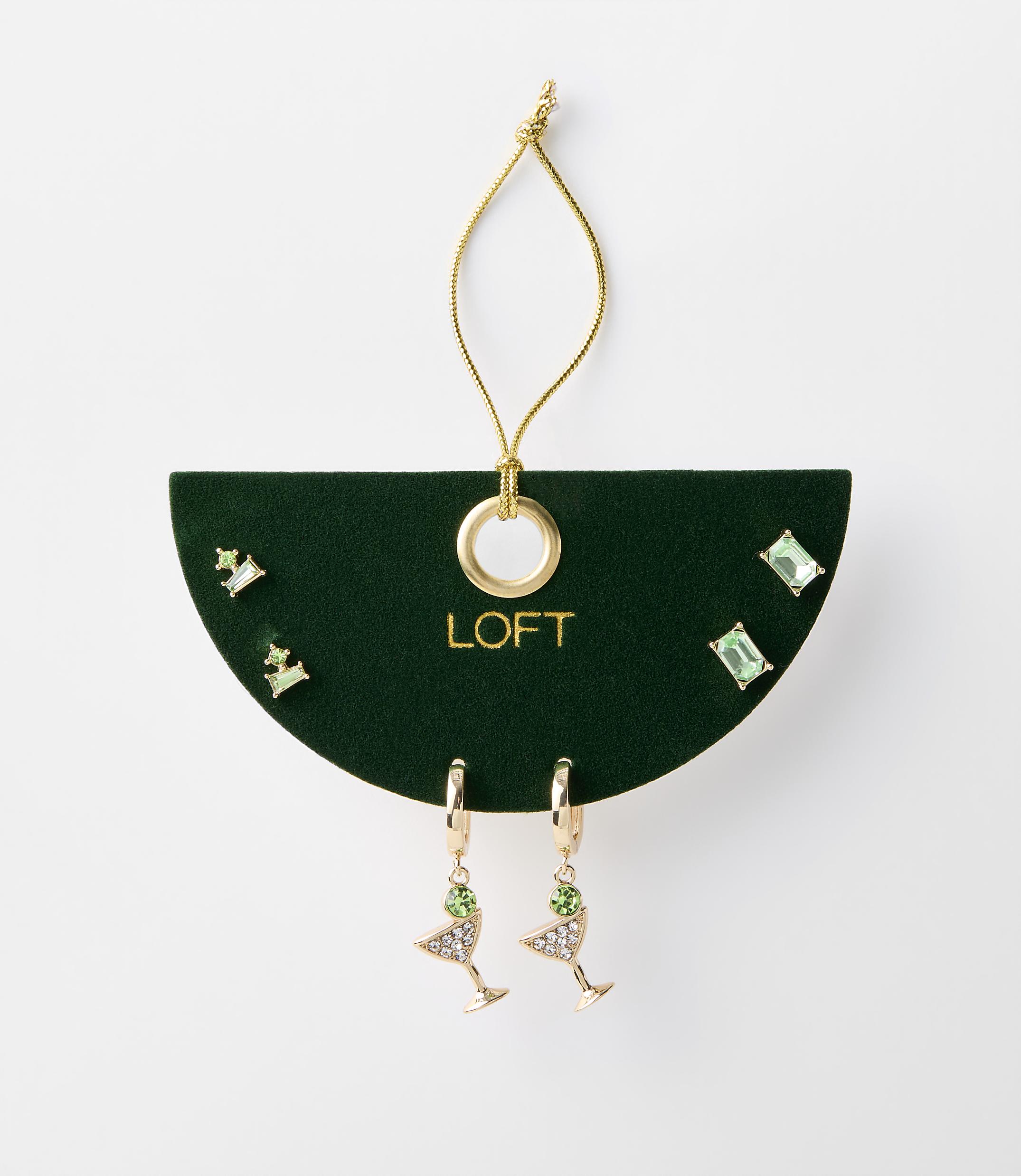 A set of green jeweled earrings, including martini glass and stud styles, are displayed on a dark green card with a gold loop and string against a plain white background.