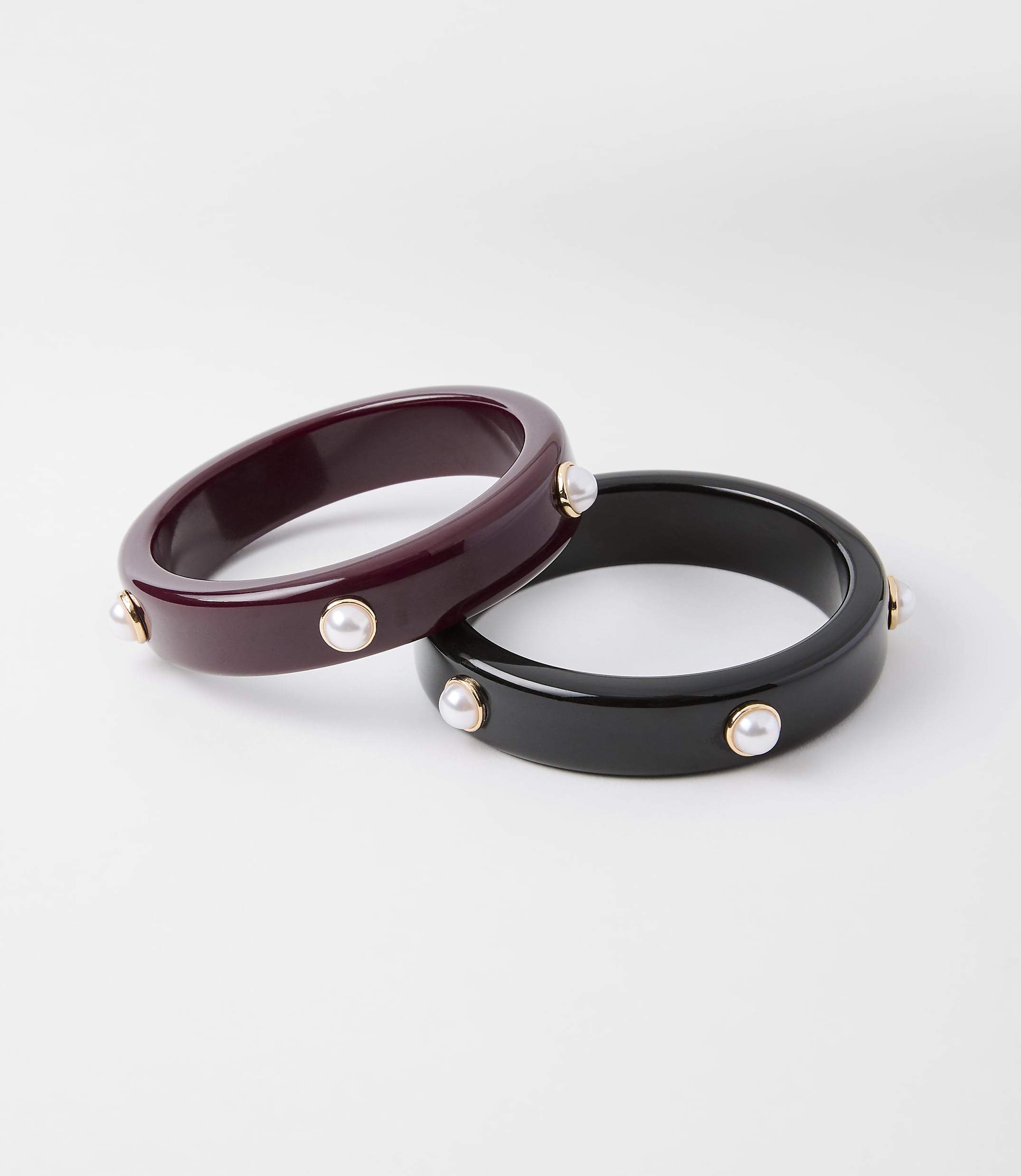 Two chunky bangles, one deep red and the other black, are adorned with small pearls and gold accents, set against a plain white background.