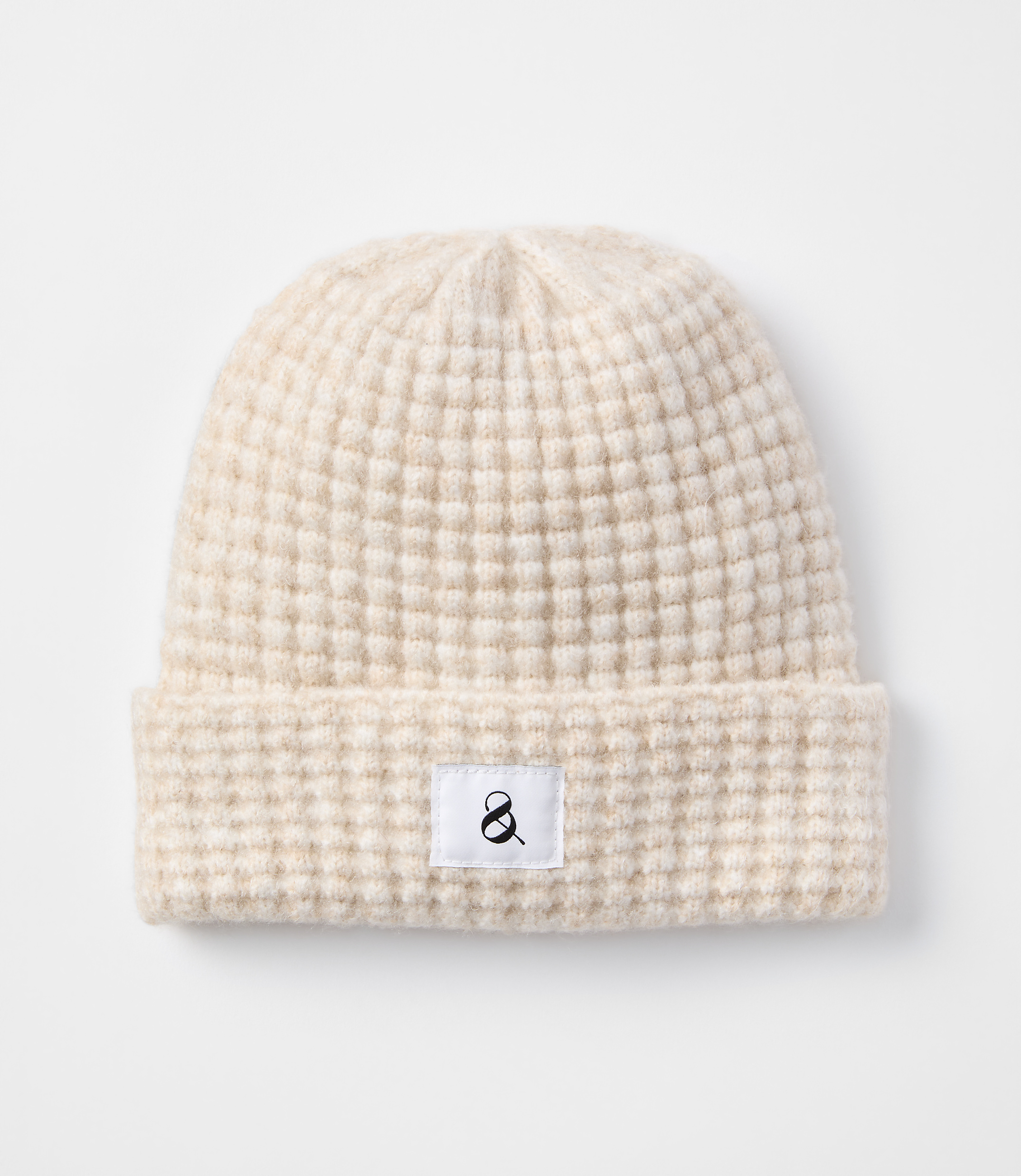 A cream-colored waffle-knit beanie with a small black ampersand logo on a white tag rests on a plain white background.