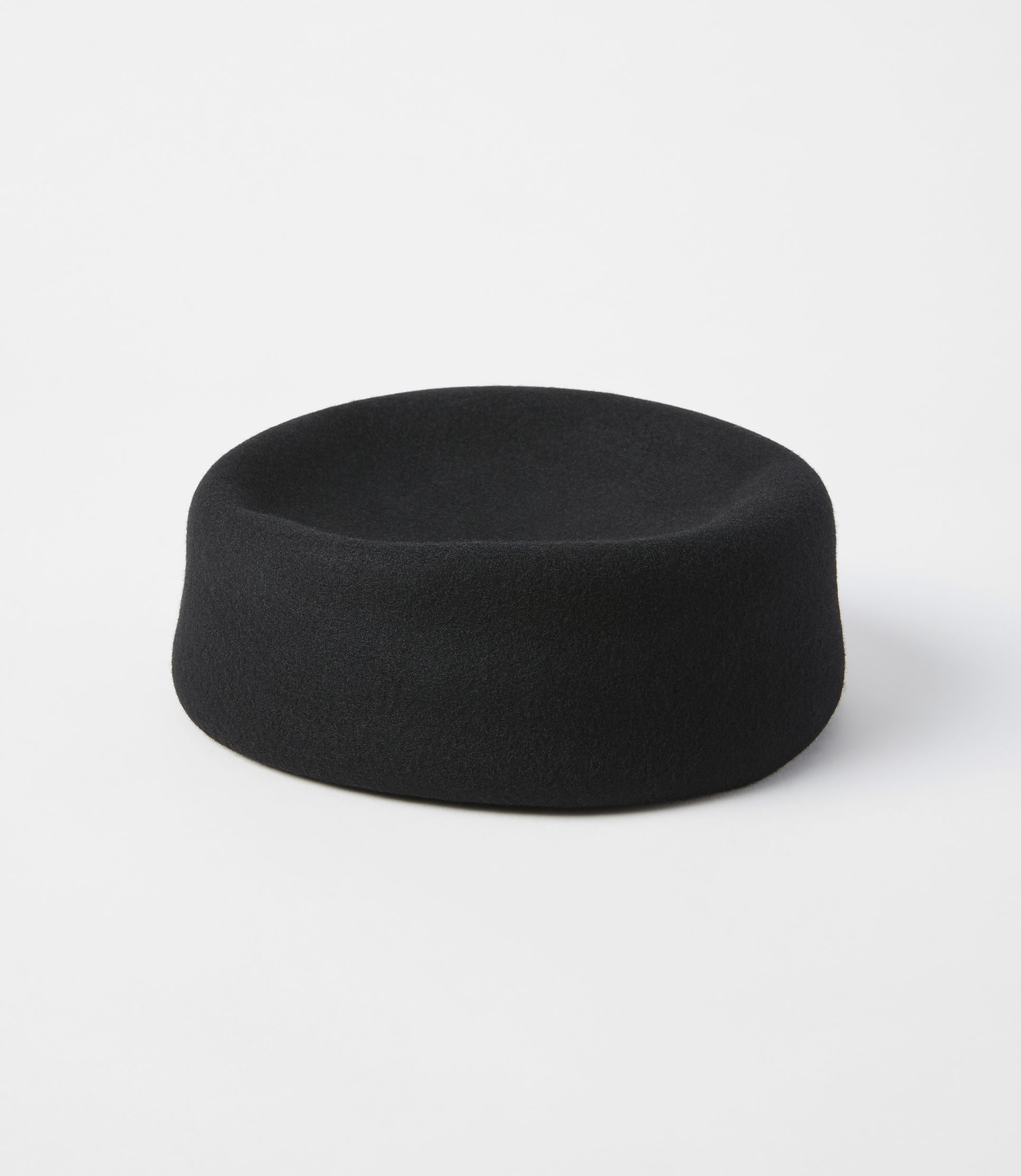 A black felt hat with a shallow crown sits on a white surface against a plain white background.