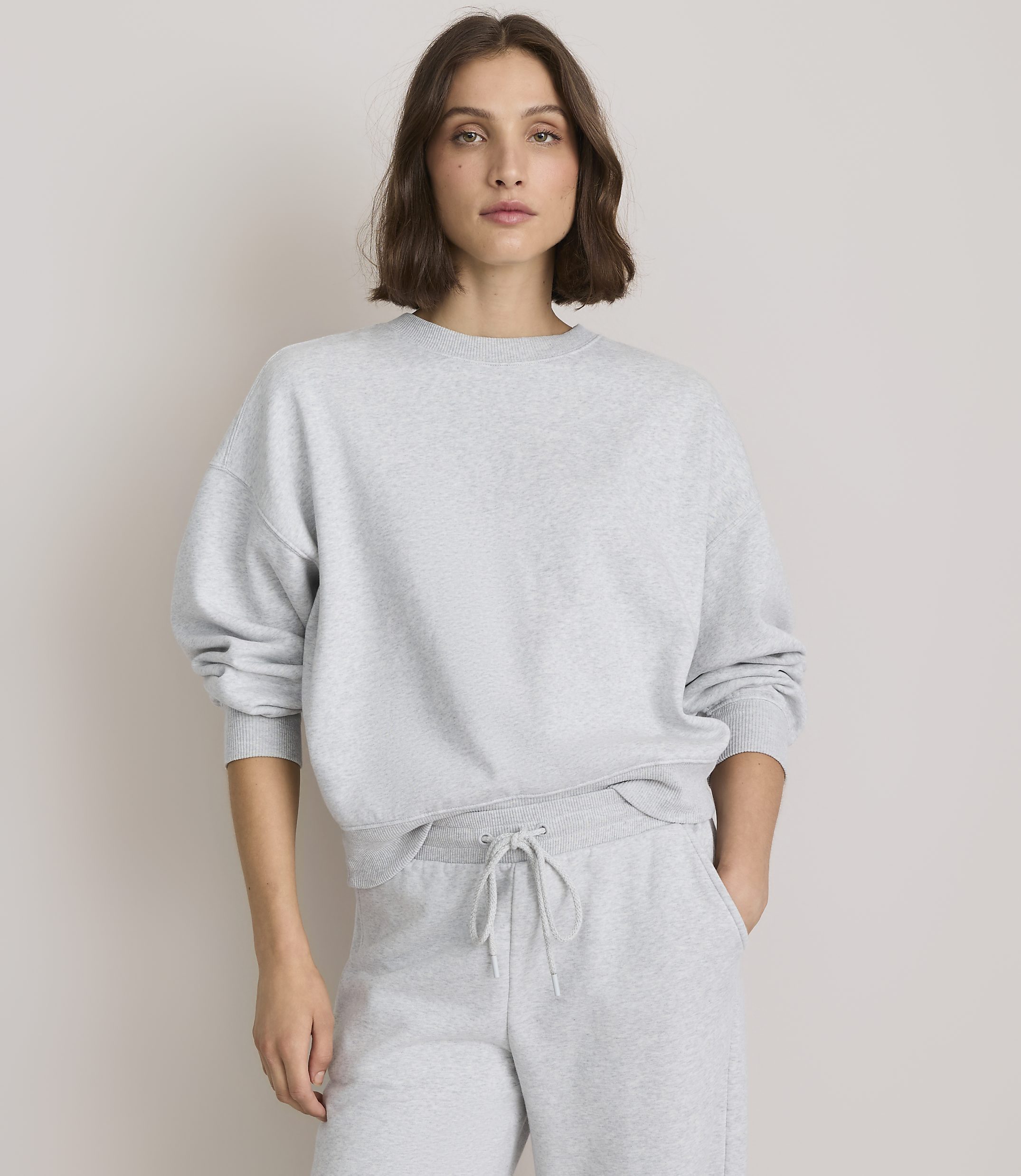 A woman in a grey sweatshirt and sweatpants stands against a plain, light grey background.