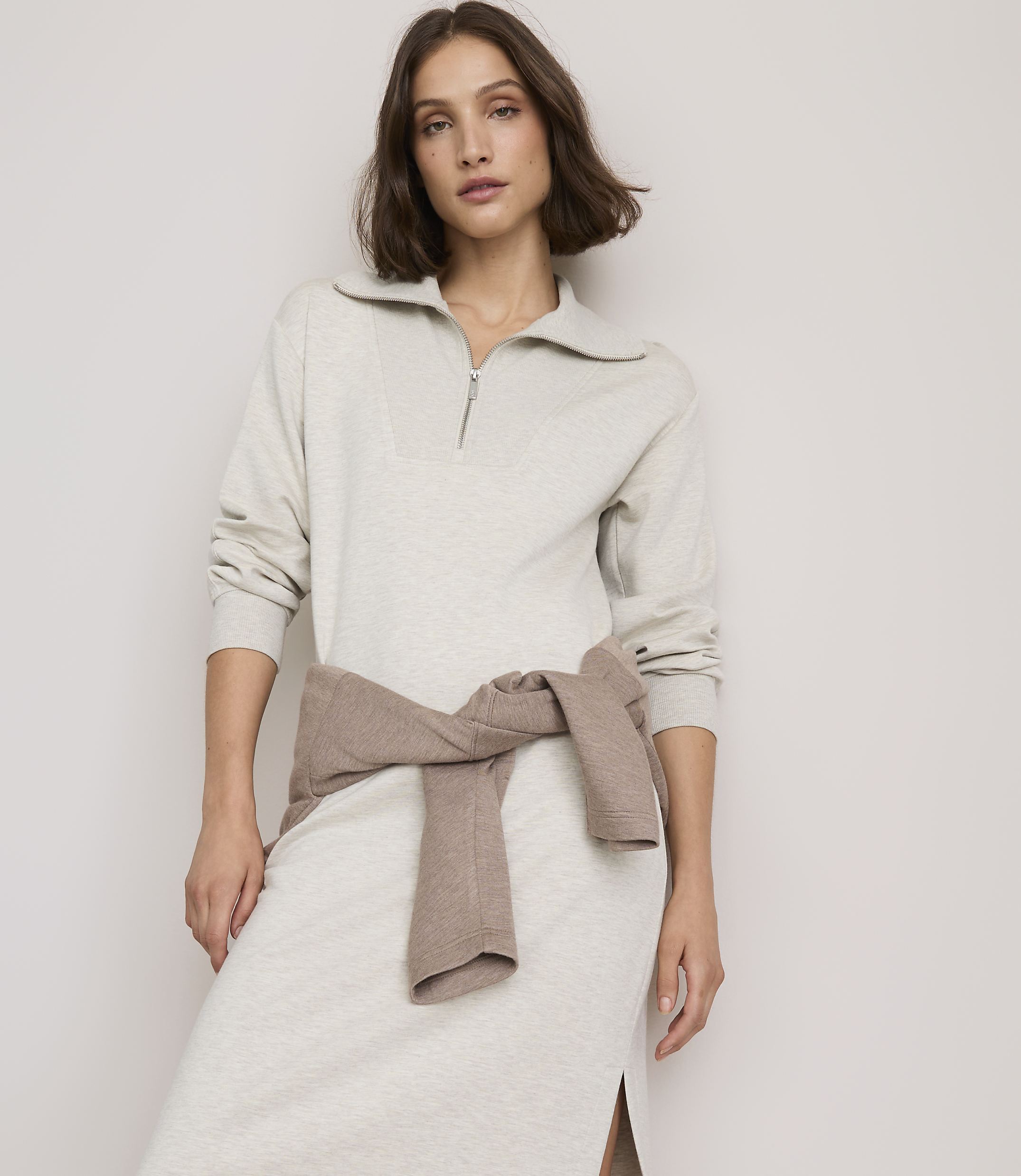 A woman in a light gray collared dress with a brown sweater tied around her waist stands against a plain, light-colored background.