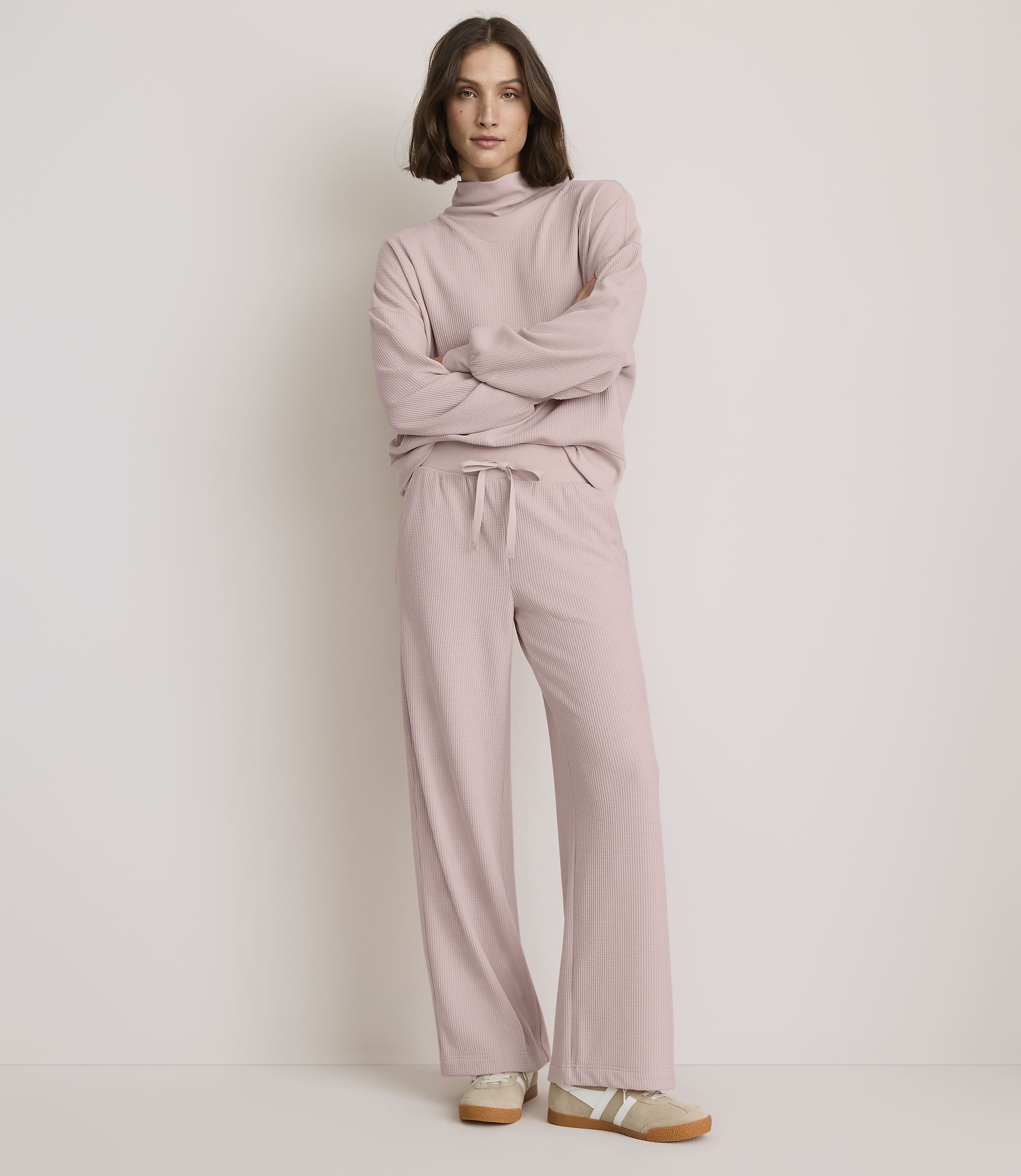 A woman in a pink ribbed loungewear set stands with her arms crossed against a plain, light-colored background.