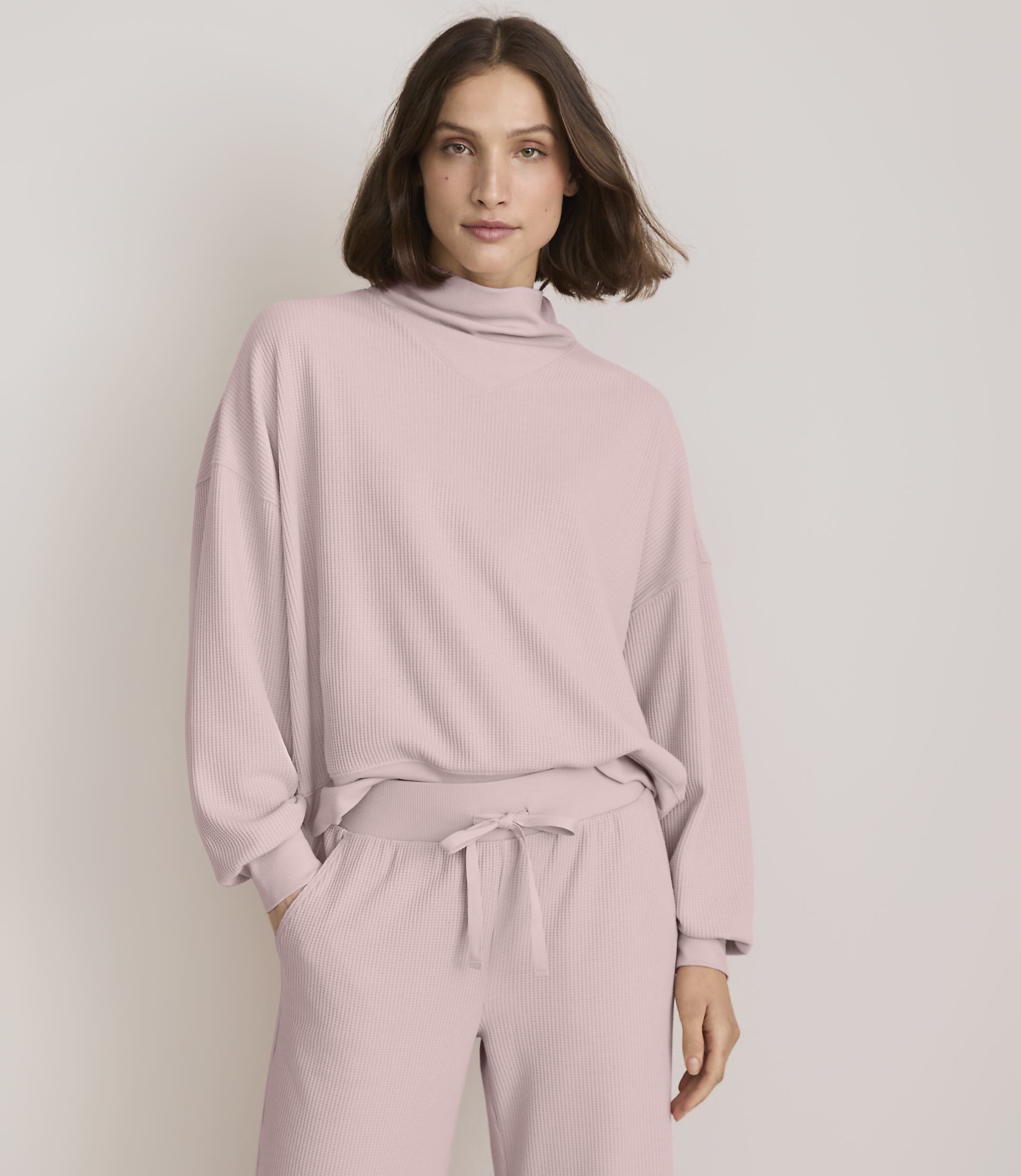 A woman in a pink ribbed turtleneck sweater and matching pants stands against a plain white background.