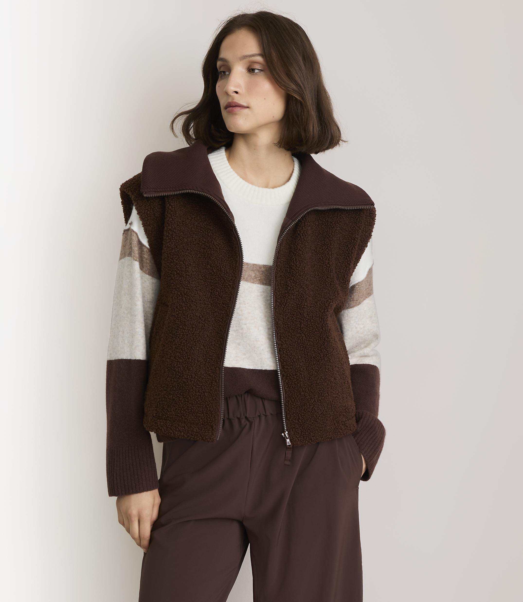 A woman in a brown fuzzy vest and striped sweater stands against a plain white background.