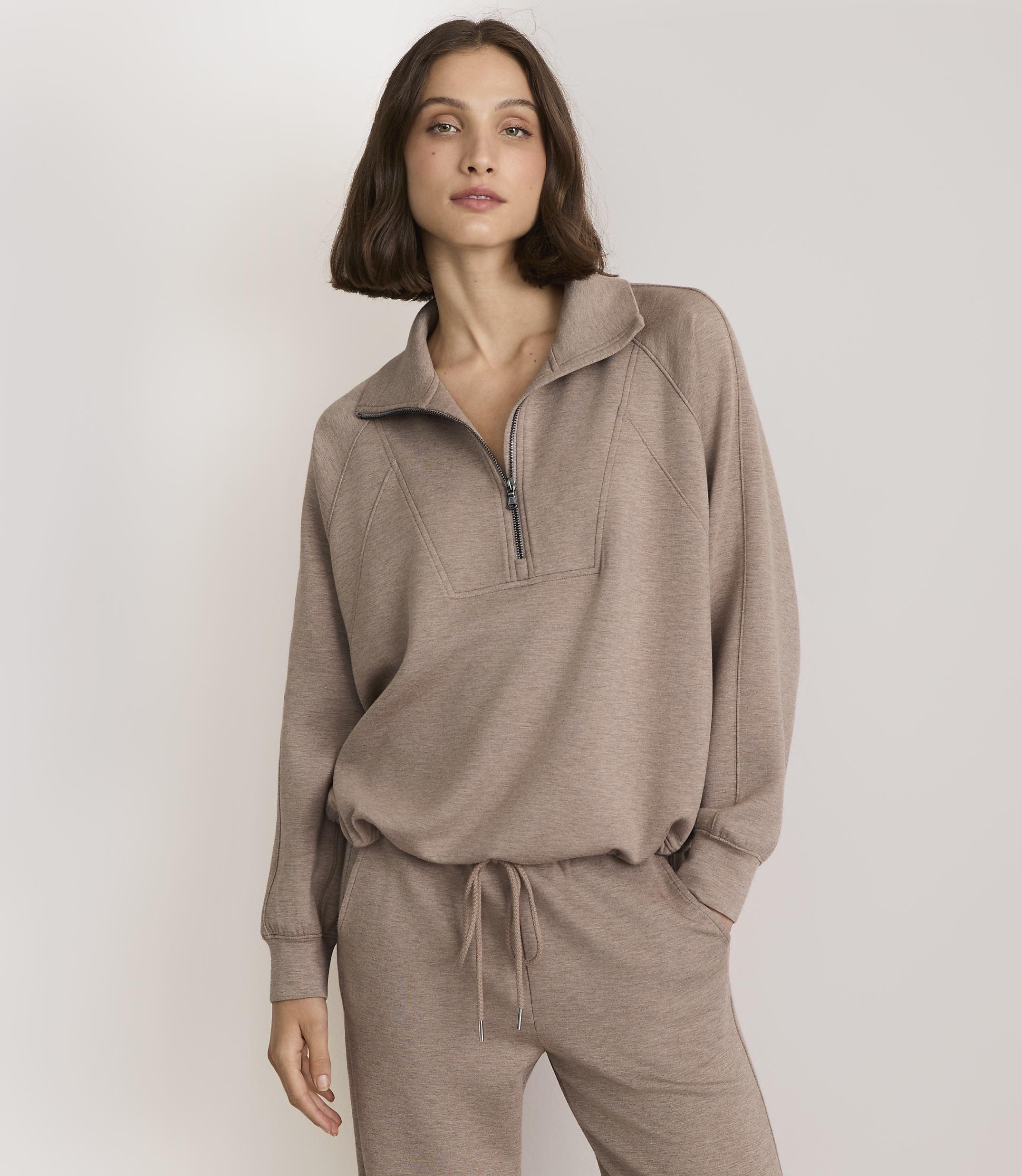 A woman in a taupe quarter-zip sweatshirt and matching sweatpants stands against a plain, light gray background.