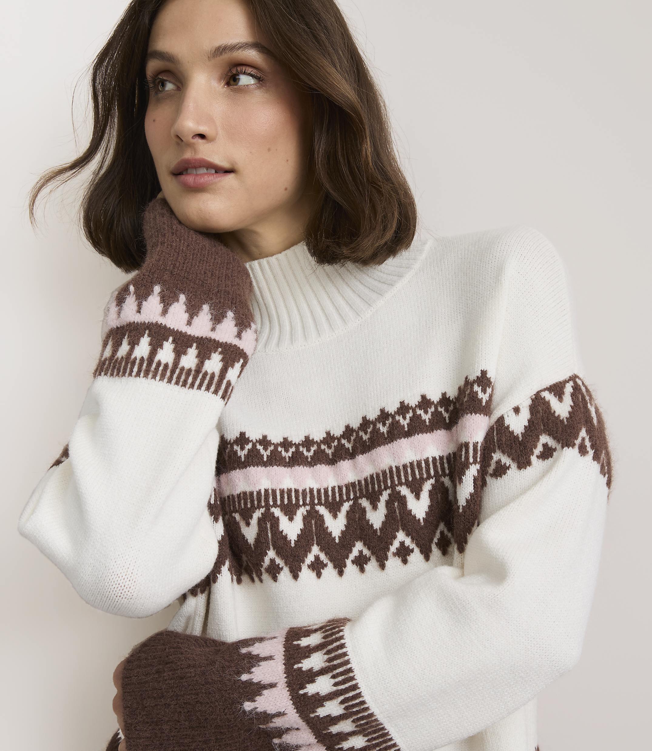 A woman in a white sweater with brown and pink geometric patterns poses against a plain white background.