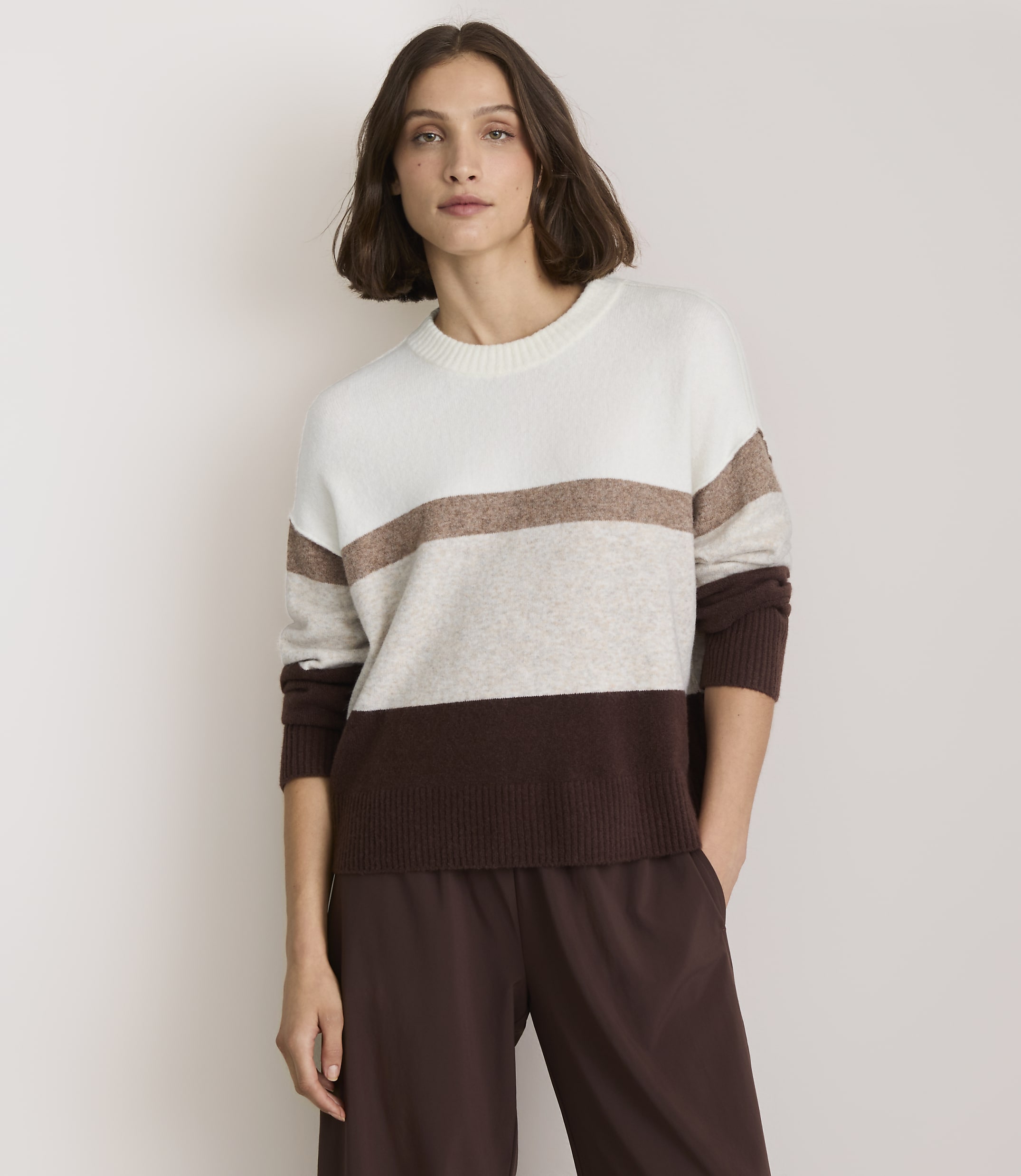 A woman in a striped sweater and brown pants stands against a plain white background.