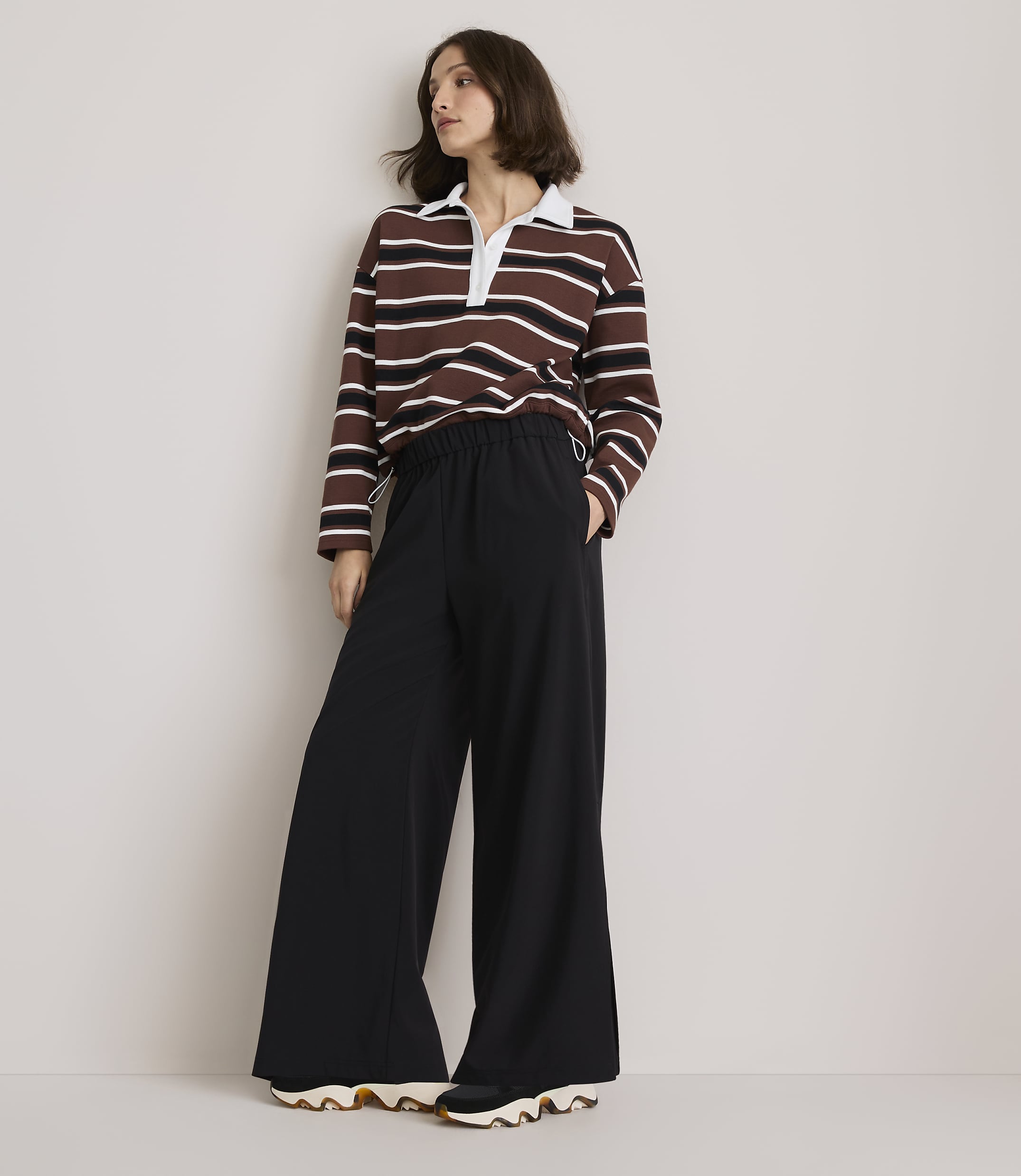A woman in a striped rugby shirt and wide-leg black pants stands against a plain, light-colored wall.