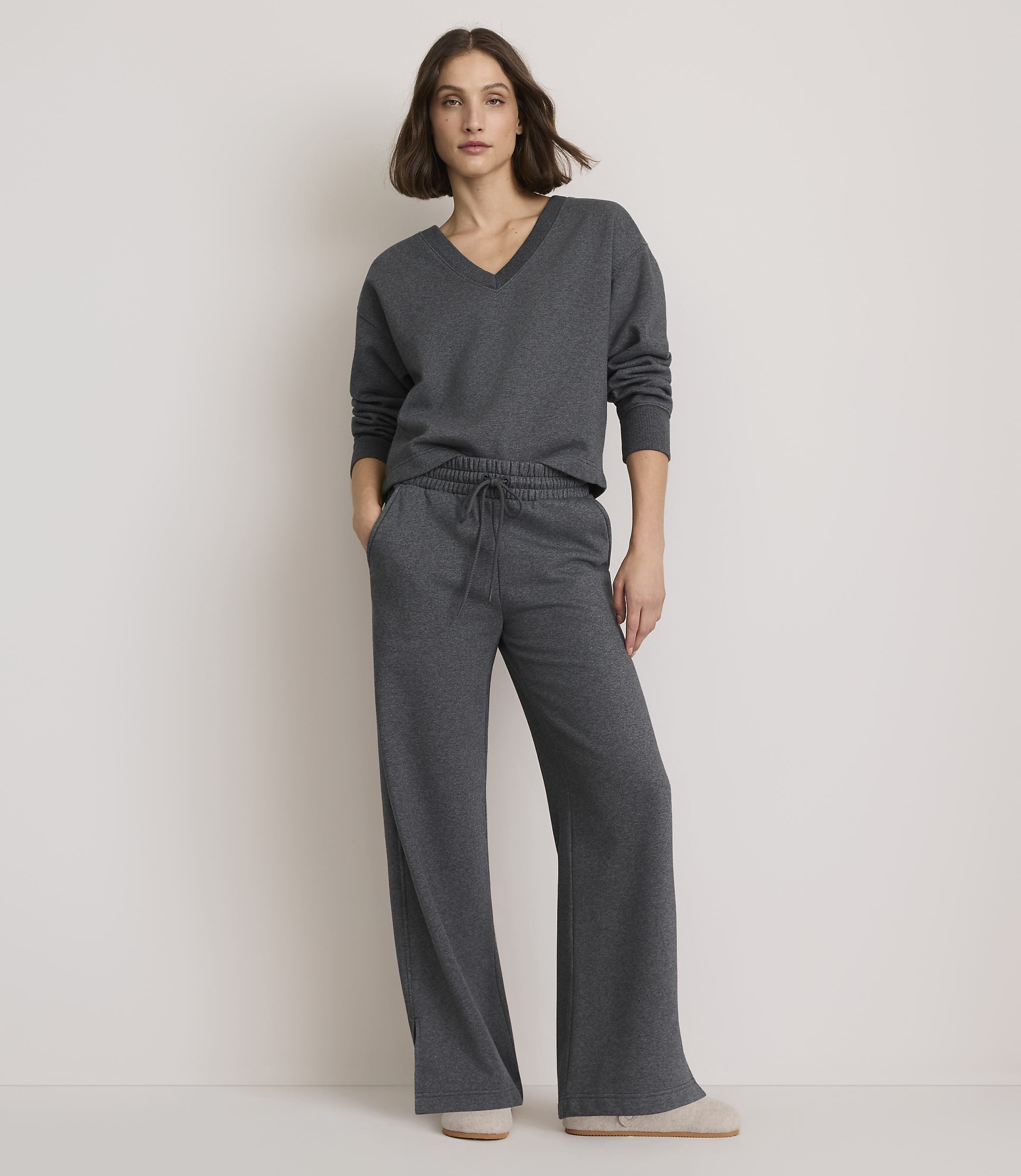 A woman in a grey v-neck sweater and wide-leg sweatpants stands against a plain white background.