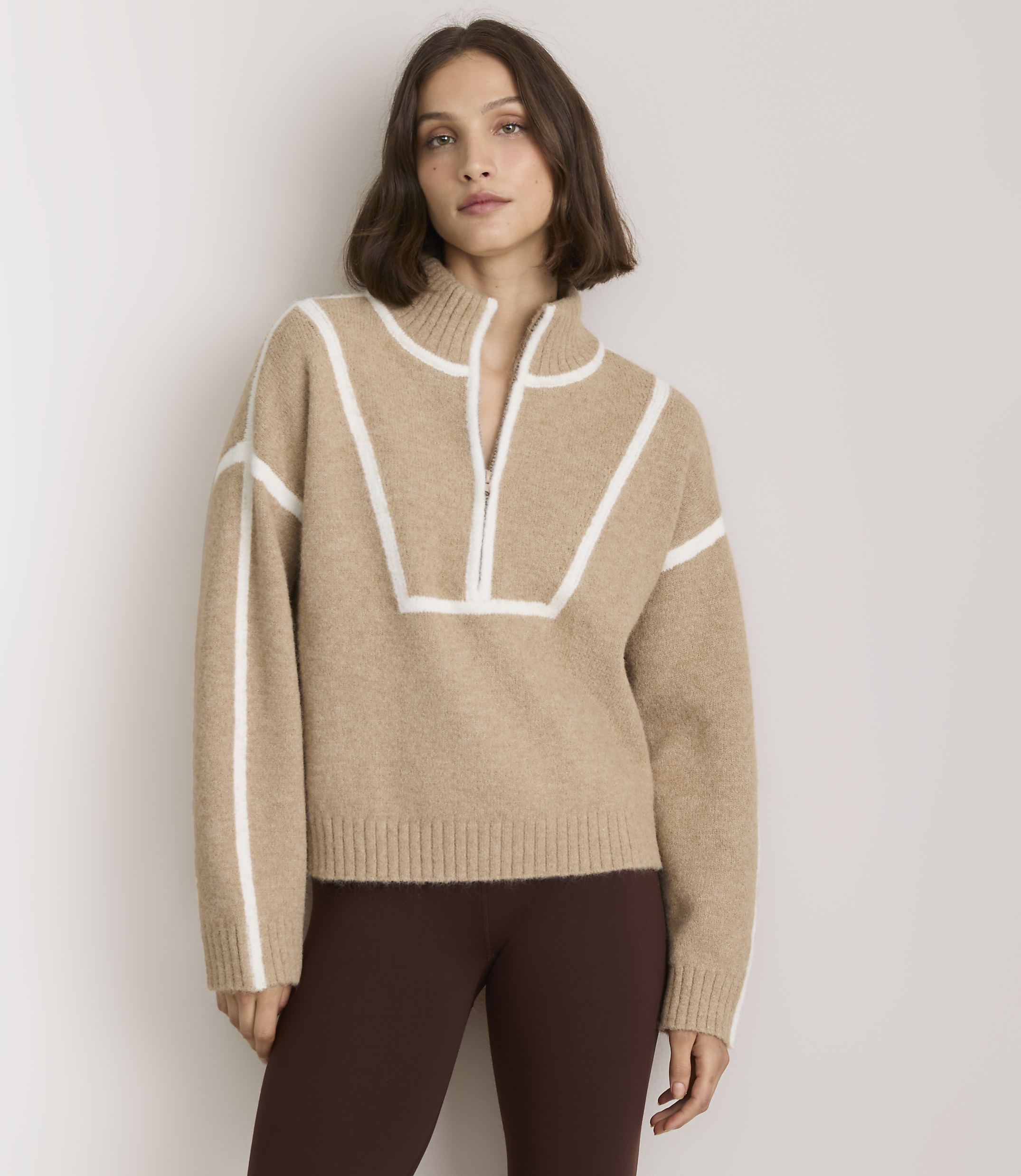 A woman in a tan quarter-zip sweater and dark leggings stands against a plain, light gray background.