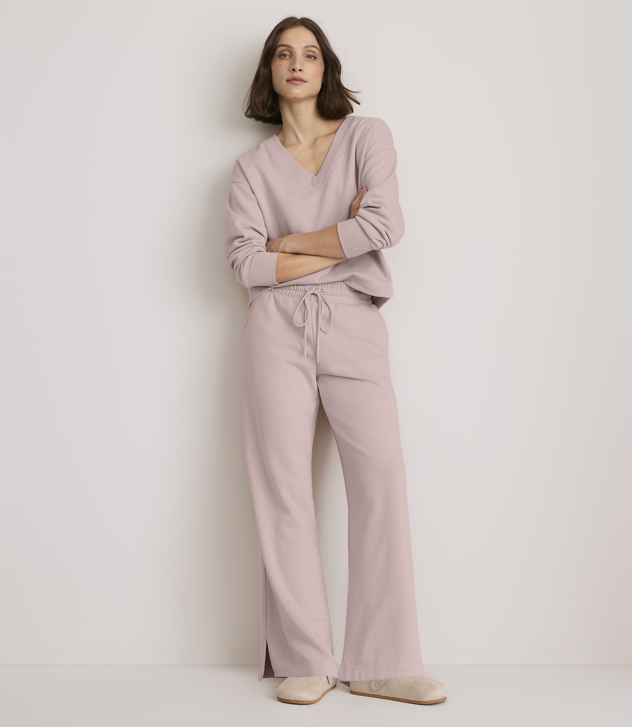 A woman in a light pink loungewear set stands with her arms crossed against a plain, light-colored wall.