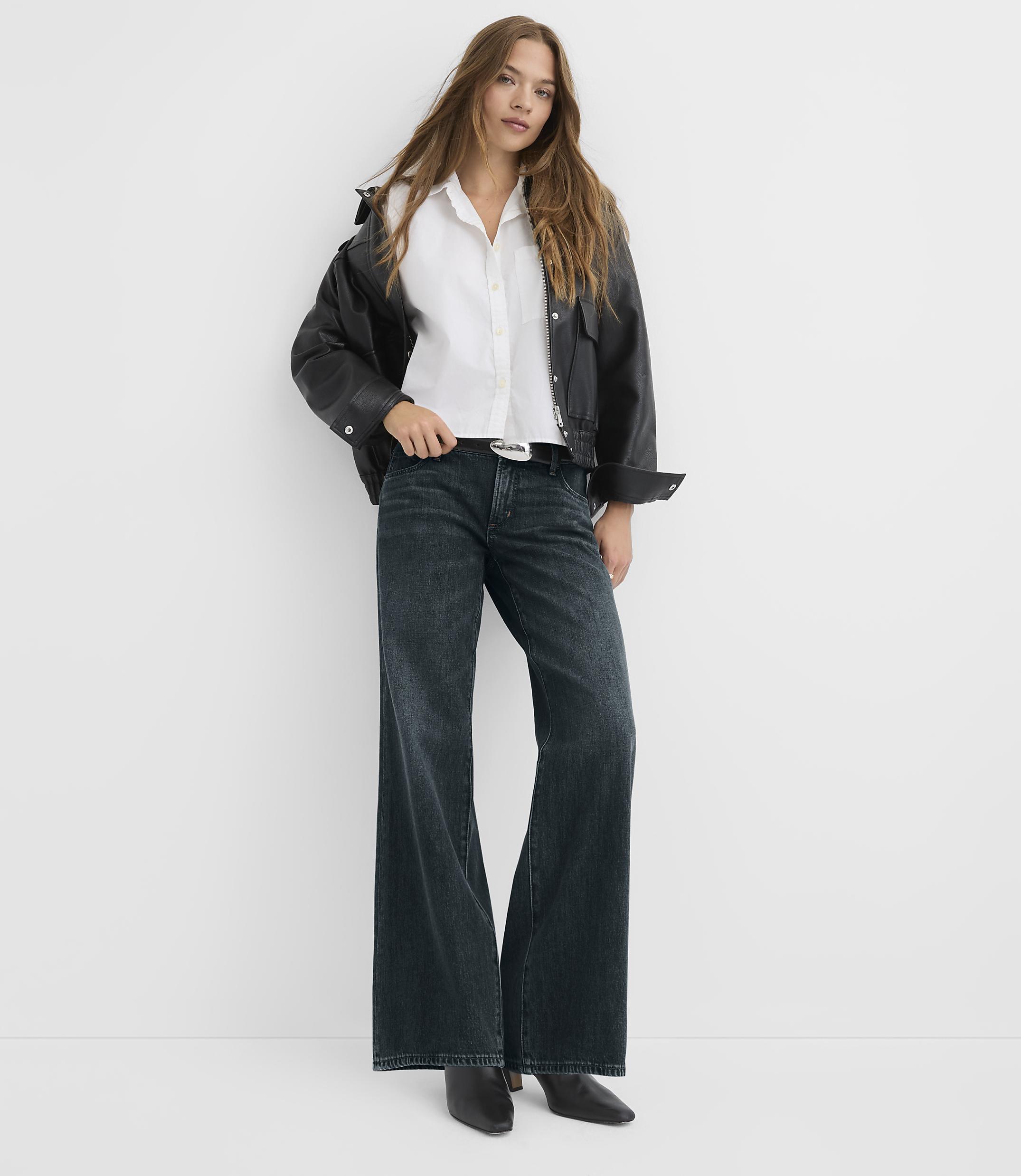 A woman in a white shirt, black jacket, and wide-leg jeans stands against a plain white background.