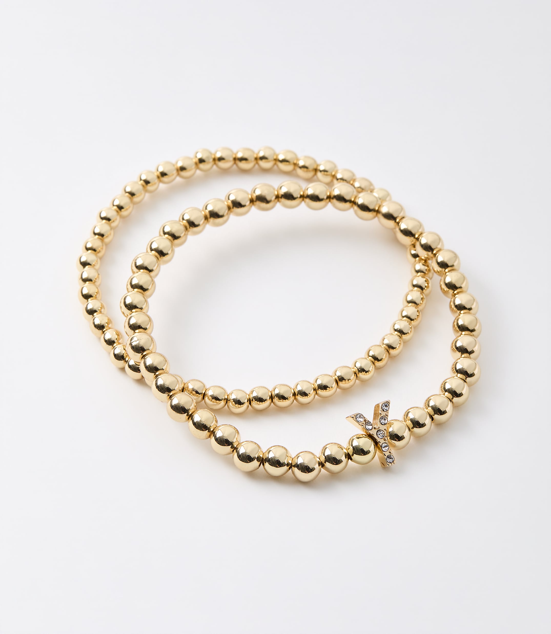 Two gold beaded bracelets, one with a diamond-studded "X" charm, are arranged on a plain white background.