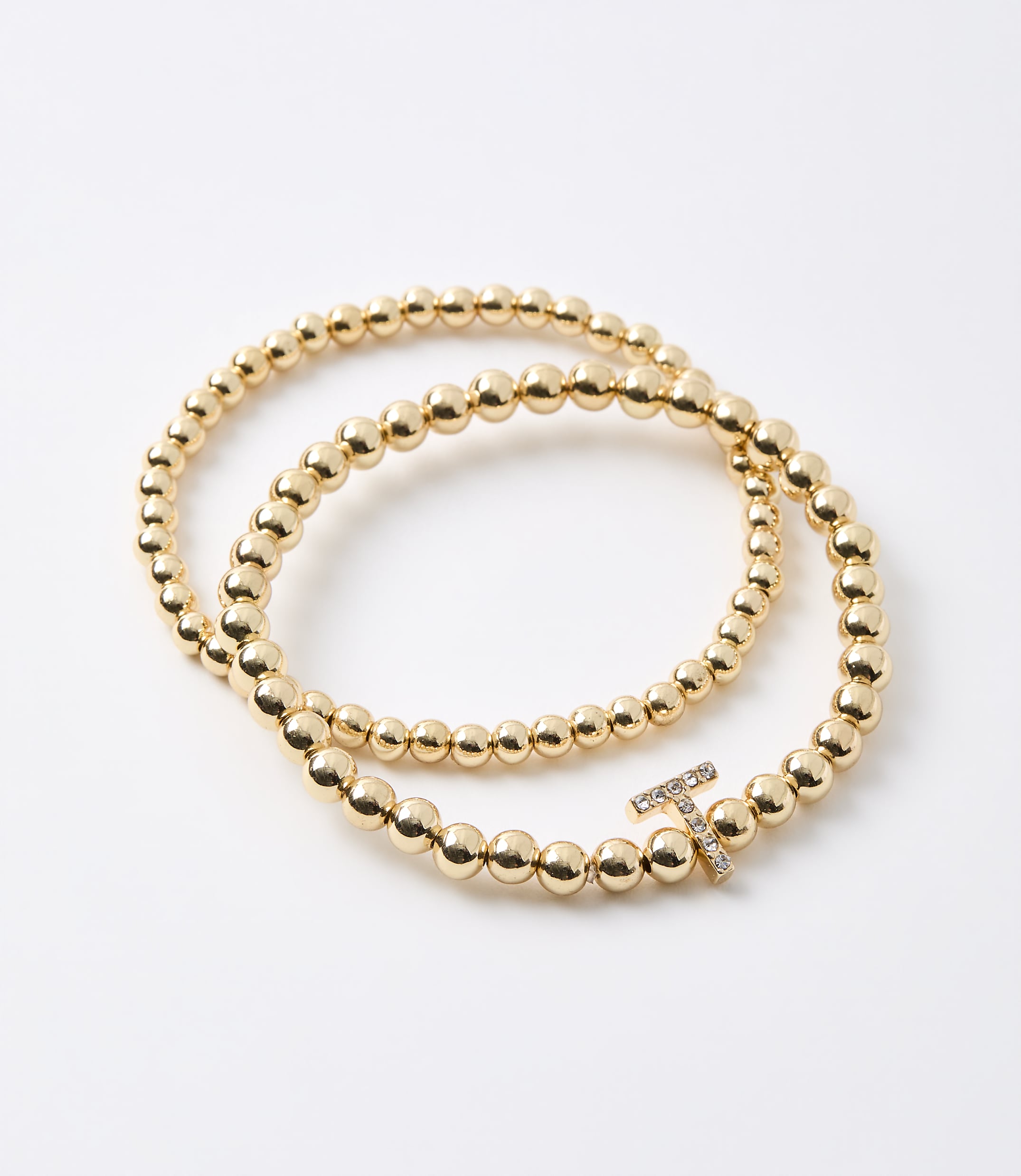 Two gold beaded bracelets, one with a diamond-encrusted "T" charm, lie on a plain white background.