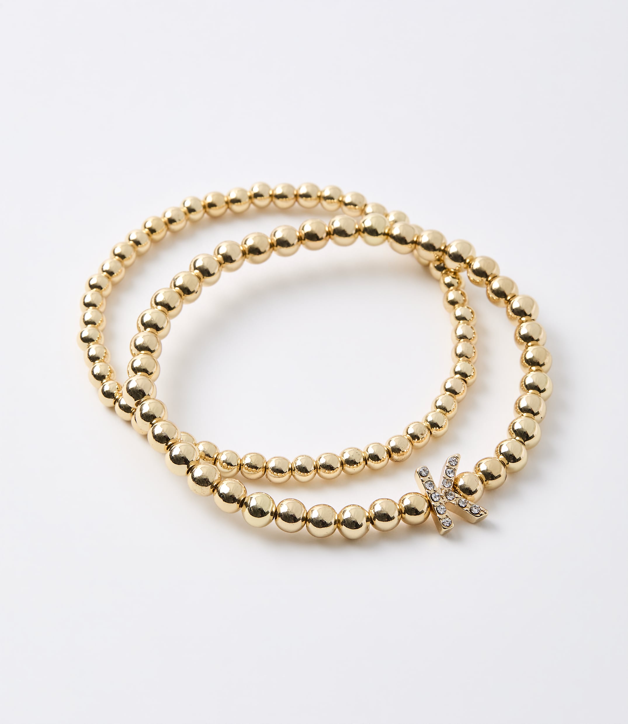 Two gold beaded bracelets, one wrapped around the other, lie on a plain white background, with one bracelet featuring a small, diamond-encrusted letter "K" charm.