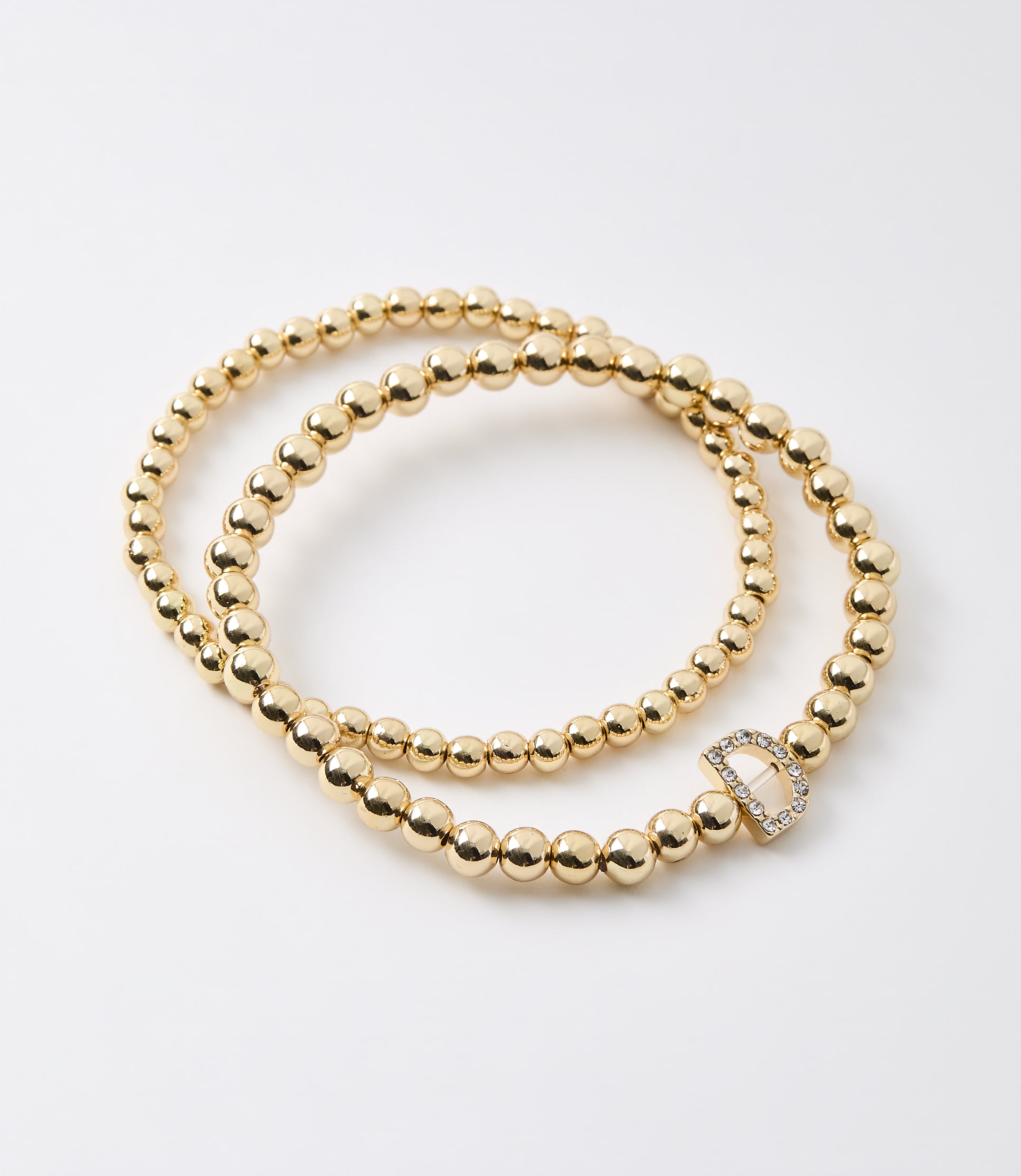 Two gold beaded bracelets, one with a diamond-encrusted "D" charm, lie on a plain white background.