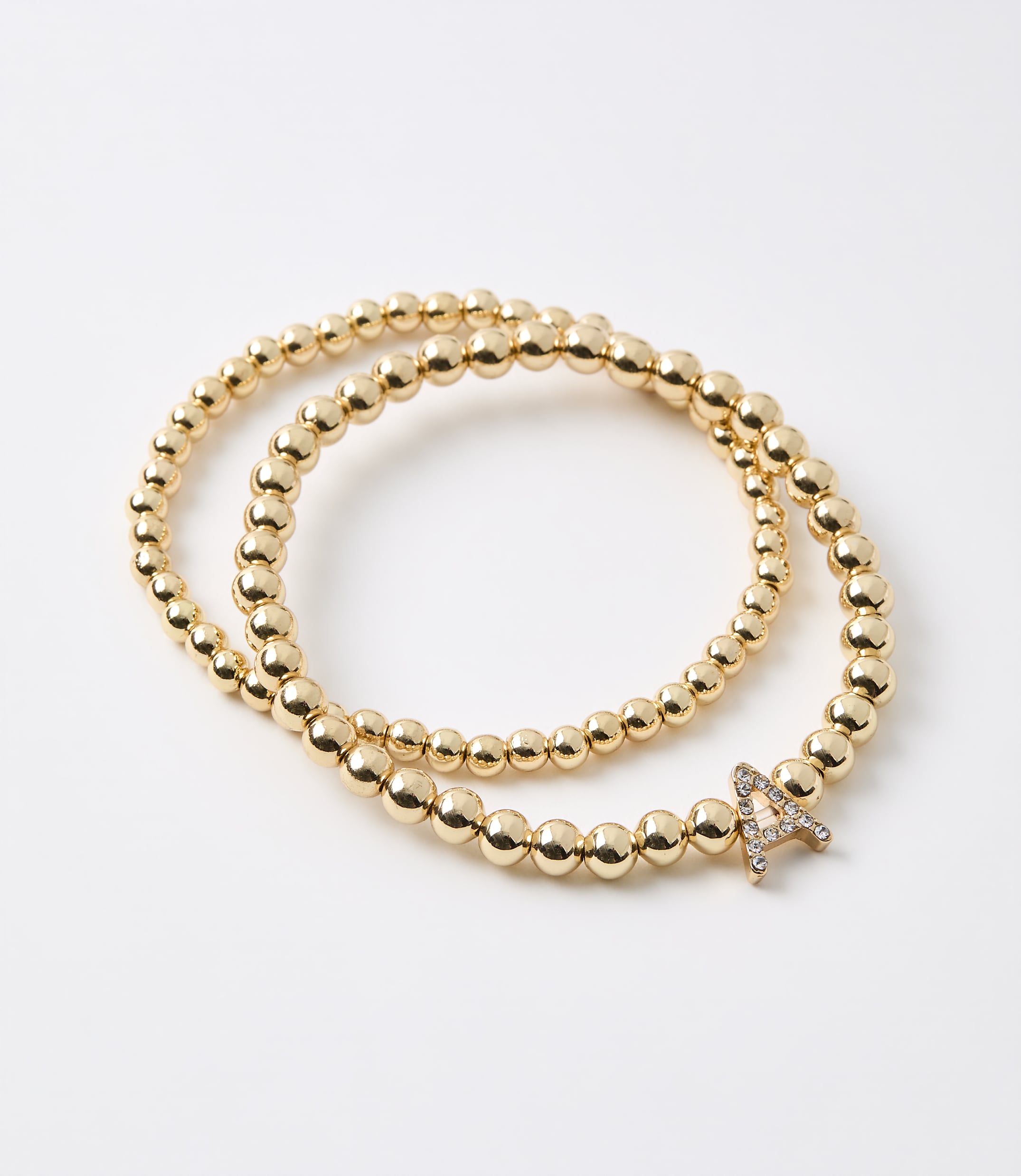 Two gold beaded bracelets, one with a diamond-encrusted letter "A" charm, lie on a plain white background.