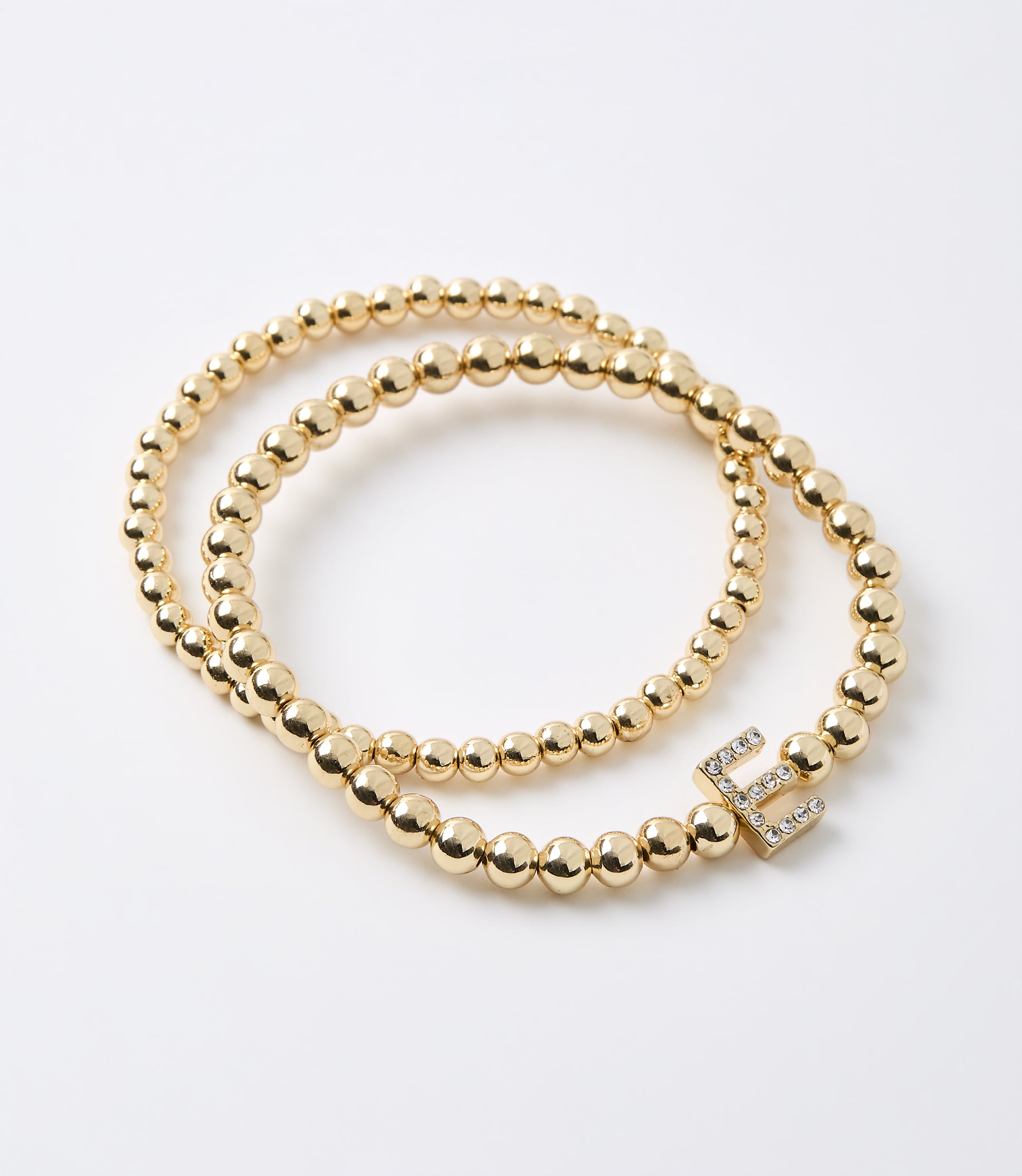 Two gold beaded bracelets, one with a diamond-encrusted letter "E" charm, lie on a plain white background.