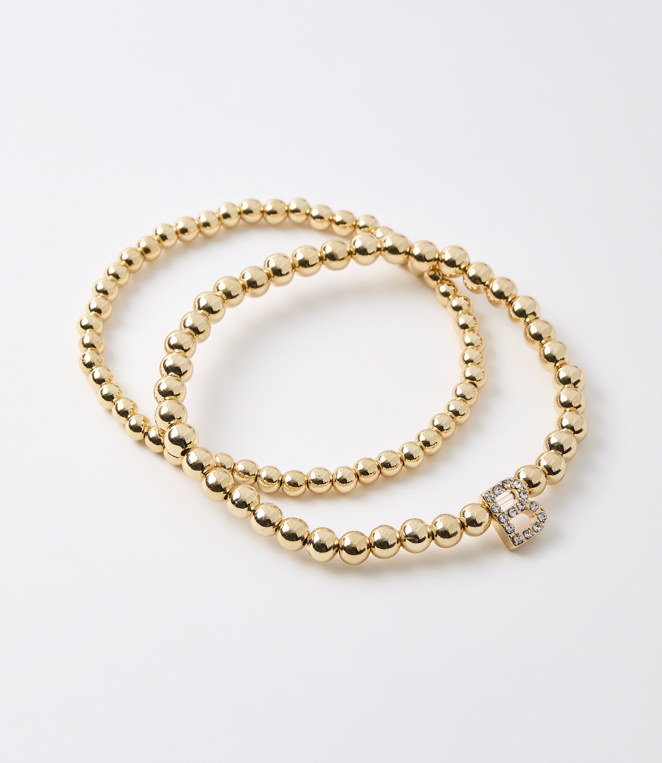 Two gold beaded bracelets, one with a diamond-encrusted letter "B" charm, lie on a plain white background.