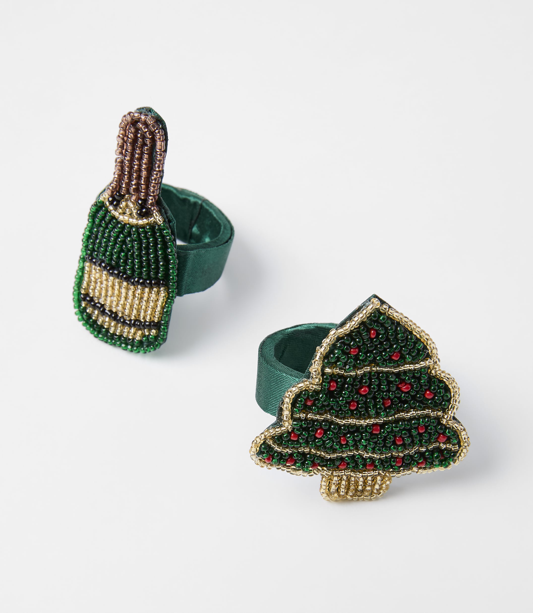 Two beaded Christmas-themed napkin rings, one shaped like a champagne bottle and the other like a Christmas tree, rest on a plain white background.