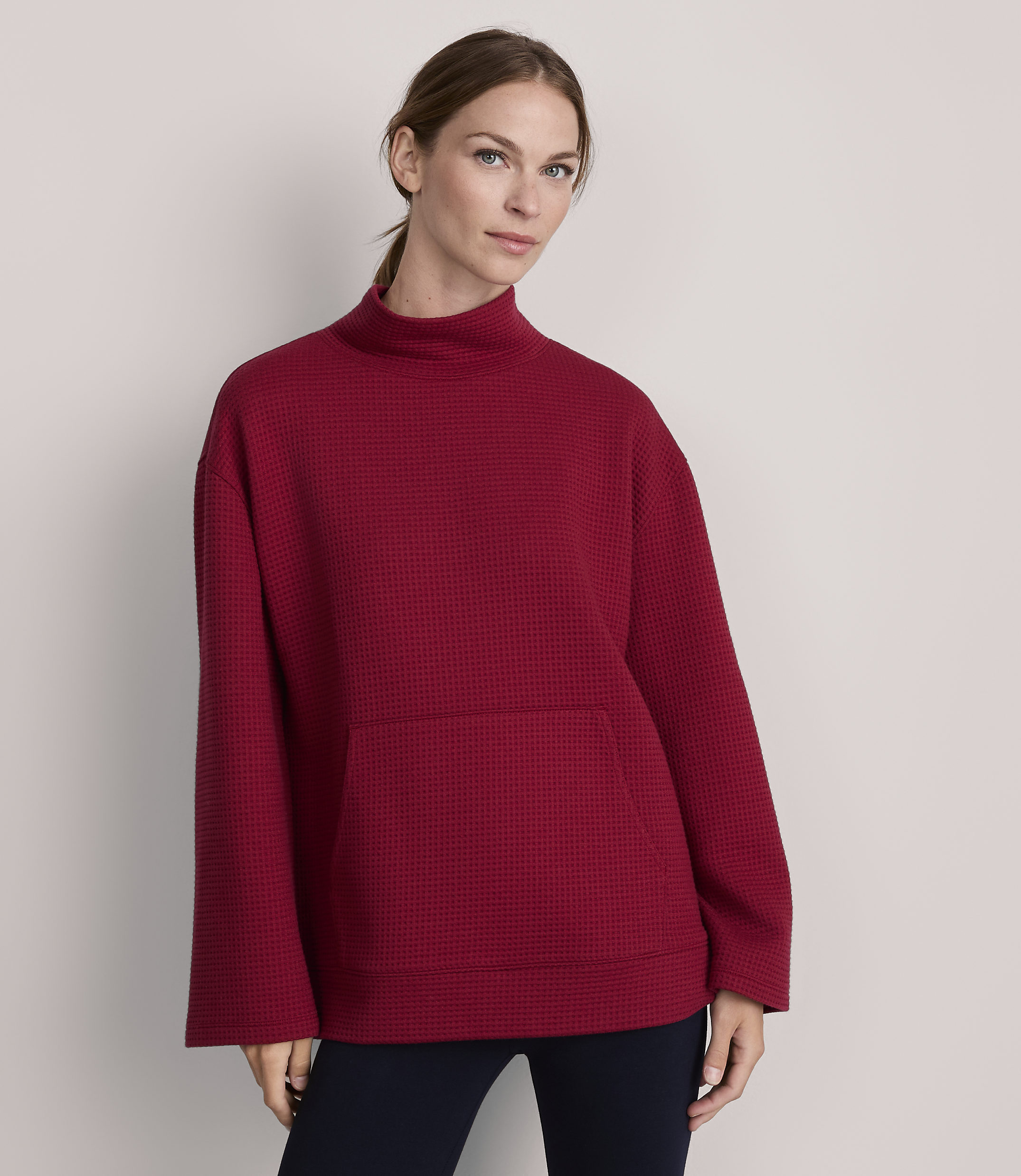 A woman in a red waffle-knit turtleneck sweater with a front pocket stands against a plain gray background.