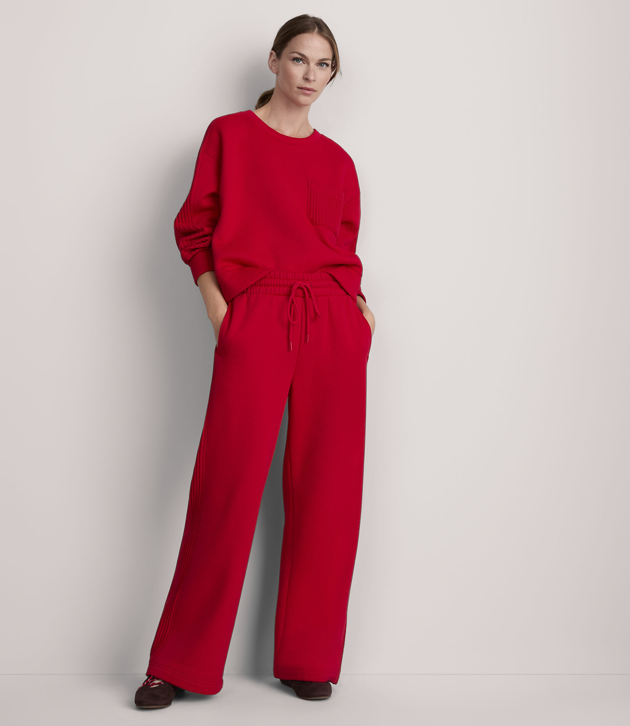A woman in a red sweatshirt and wide-leg pants stands against a plain white background.
