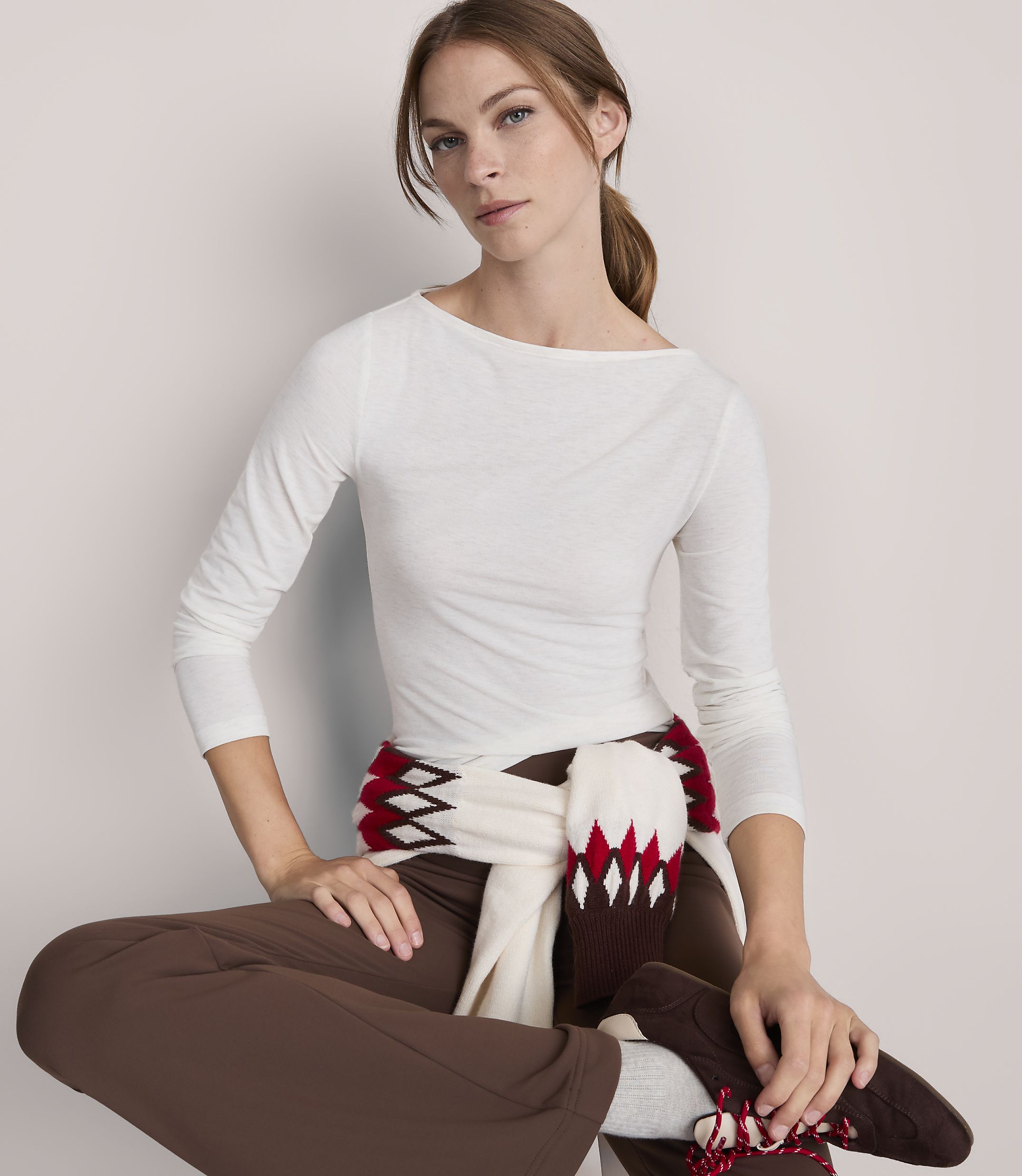 A woman in a white boatneck shirt and brown pants, with a patterned scarf draped around her waist, sits against a plain white background.
