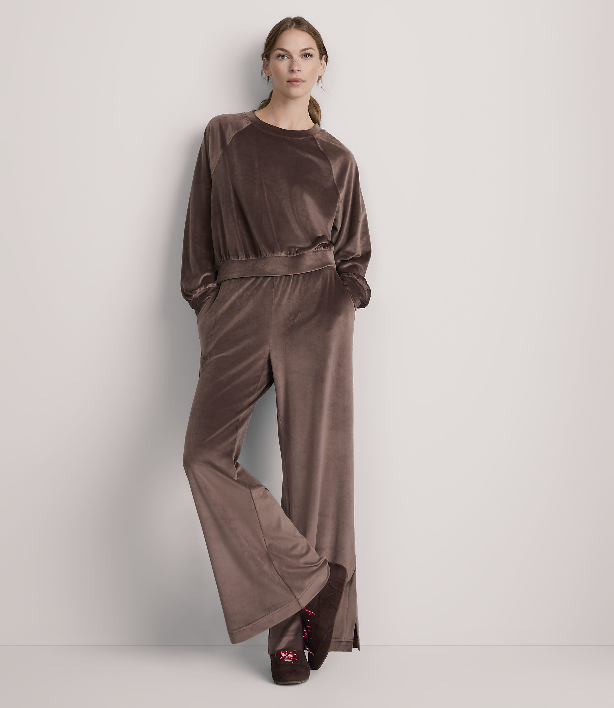 A woman in a brown velvet tracksuit stands against a plain white wall.