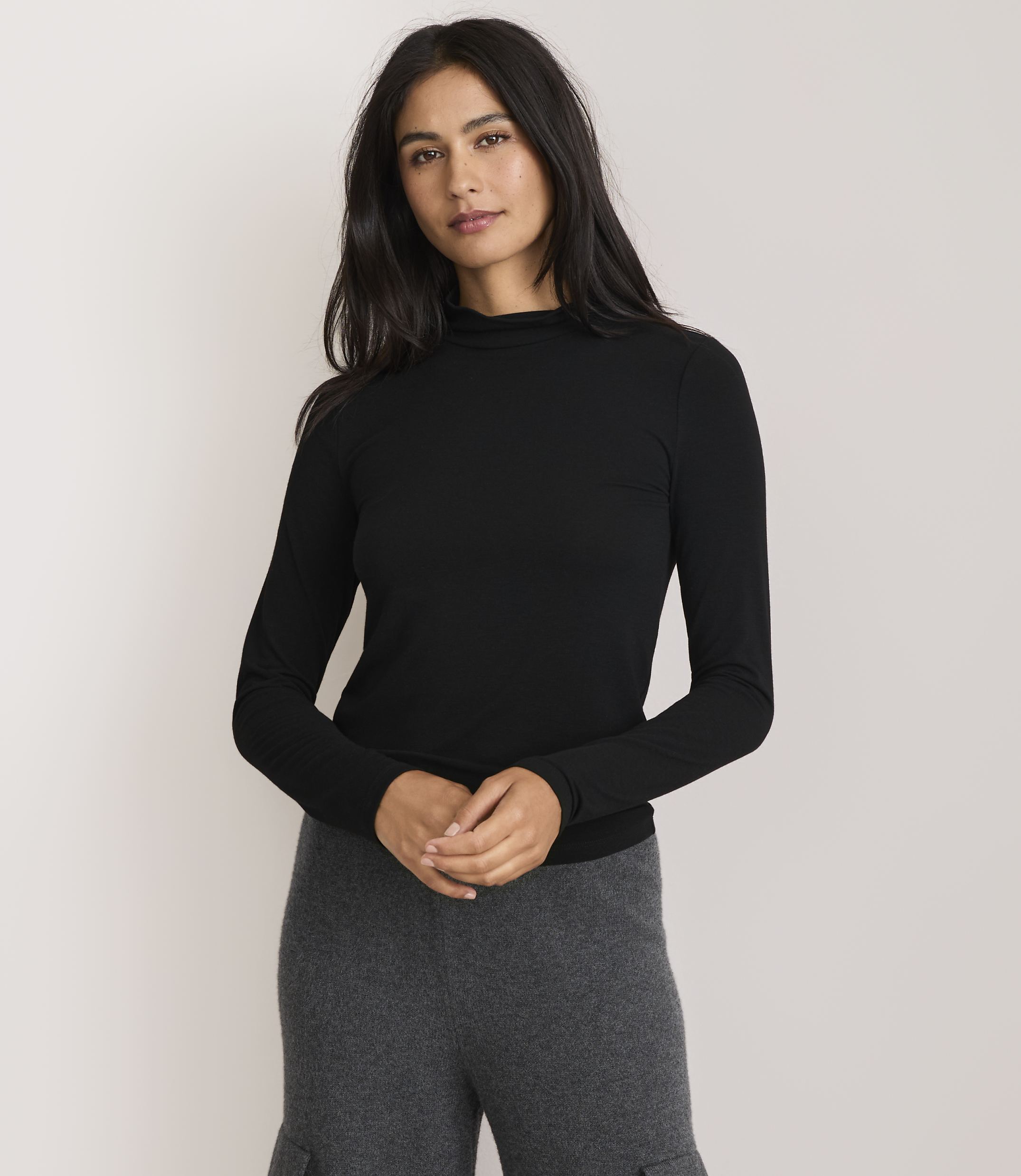 A woman in a black turtleneck and gray pants stands against a plain white background.