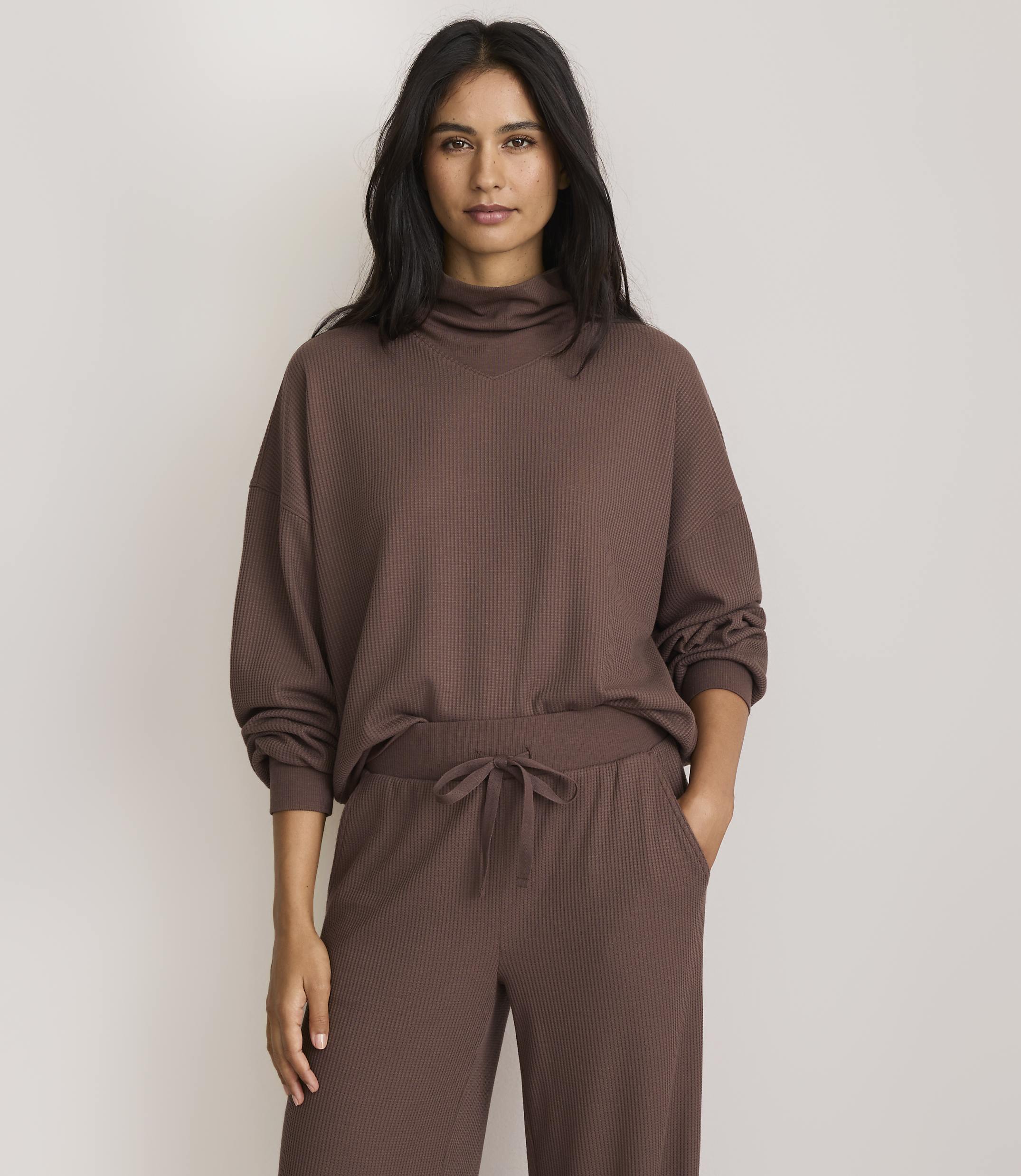 A woman in a brown turtleneck sweater and matching pants stands against a plain, light-colored background.