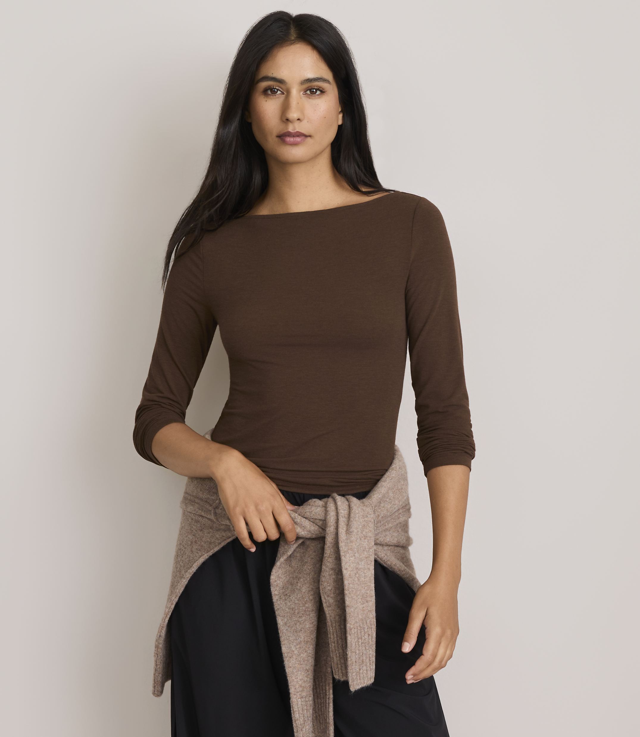 A woman in a brown boatneck shirt and black pants stands against a plain gray background, with a tan sweater tied around her waist.