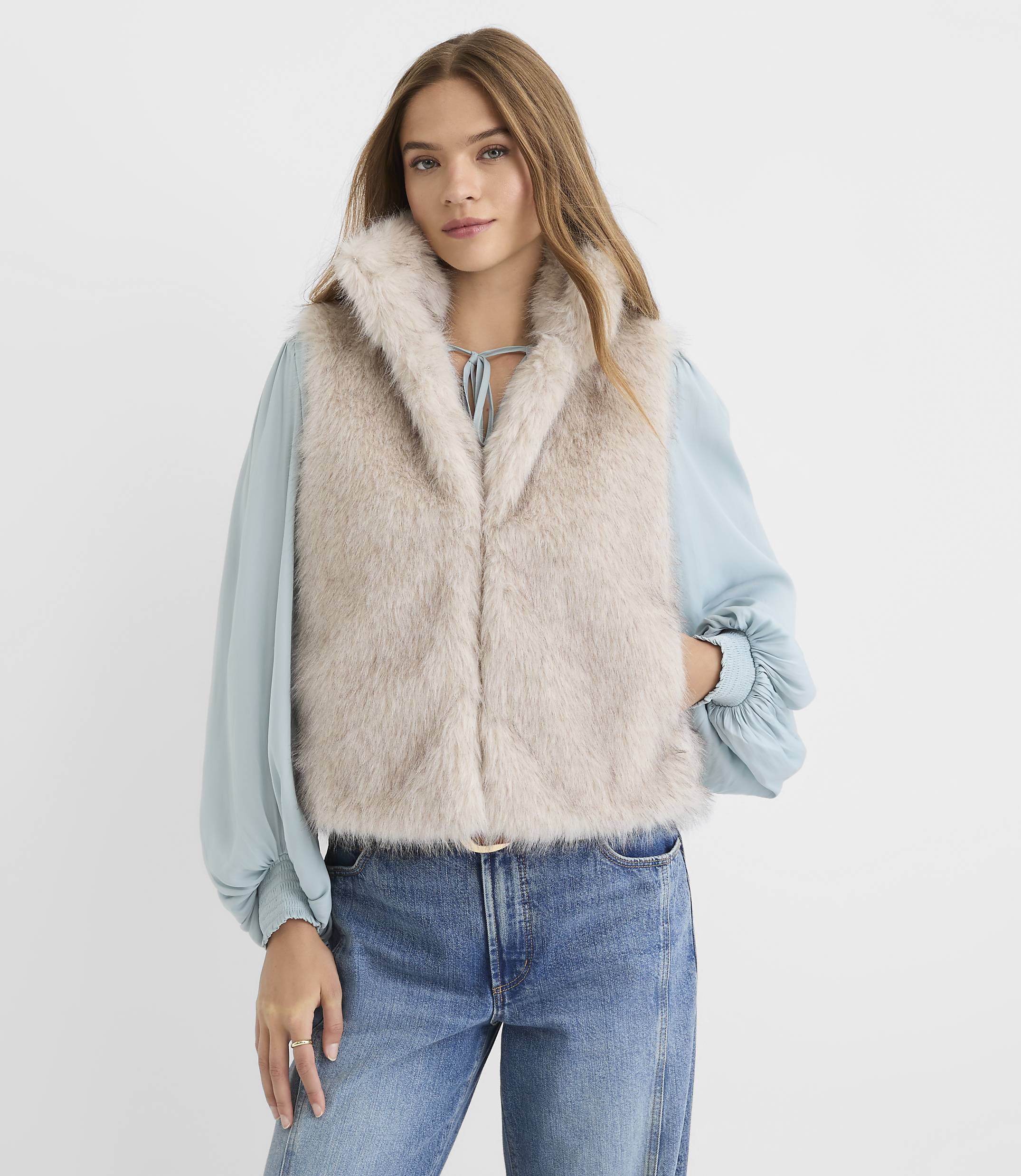 A young woman in a fuzzy vest and jeans stands against a plain, light background.