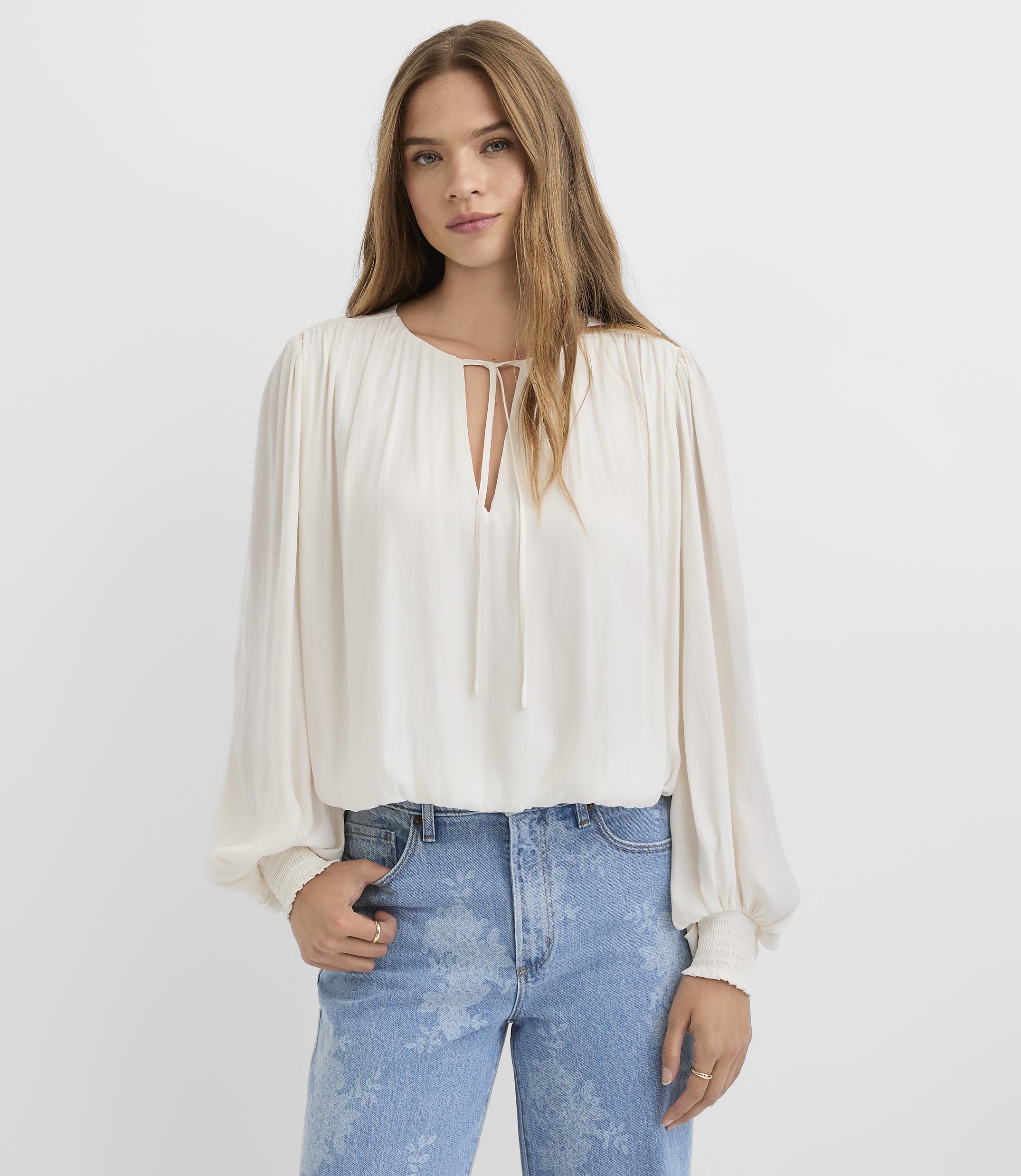 A young woman in a white bloused top and patterned jeans stands against a plain white background.