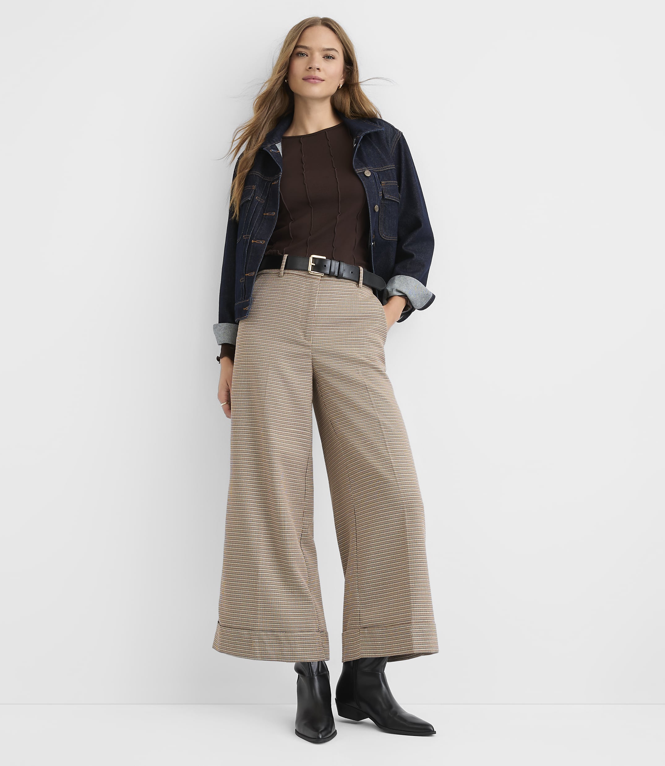 A woman in a brown ribbed top, dark denim jacket, and wide-leg plaid pants stands against a plain white background.