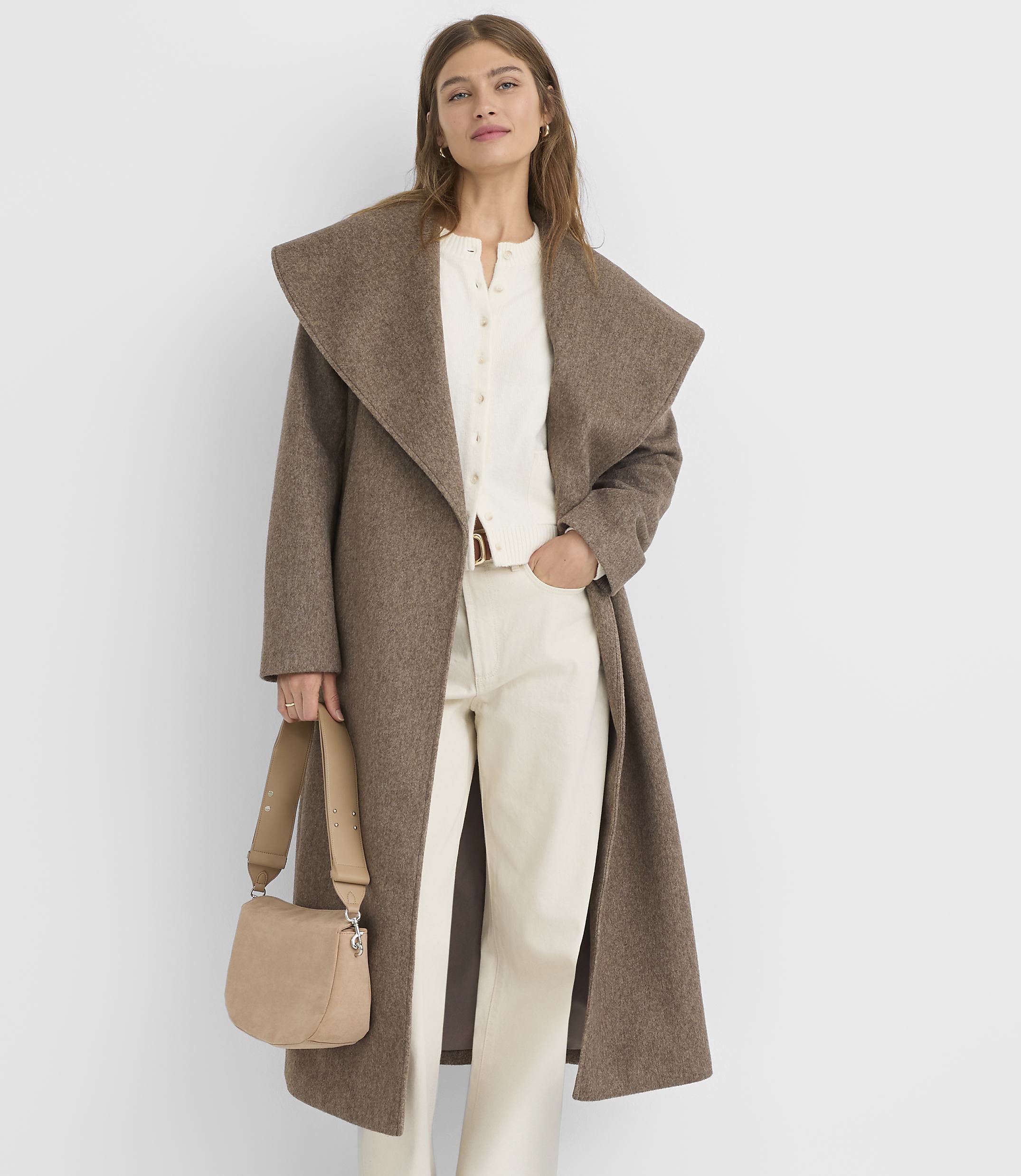 A woman in a brown coat and cream outfit stands against a plain white background, holding a beige crossbody bag.