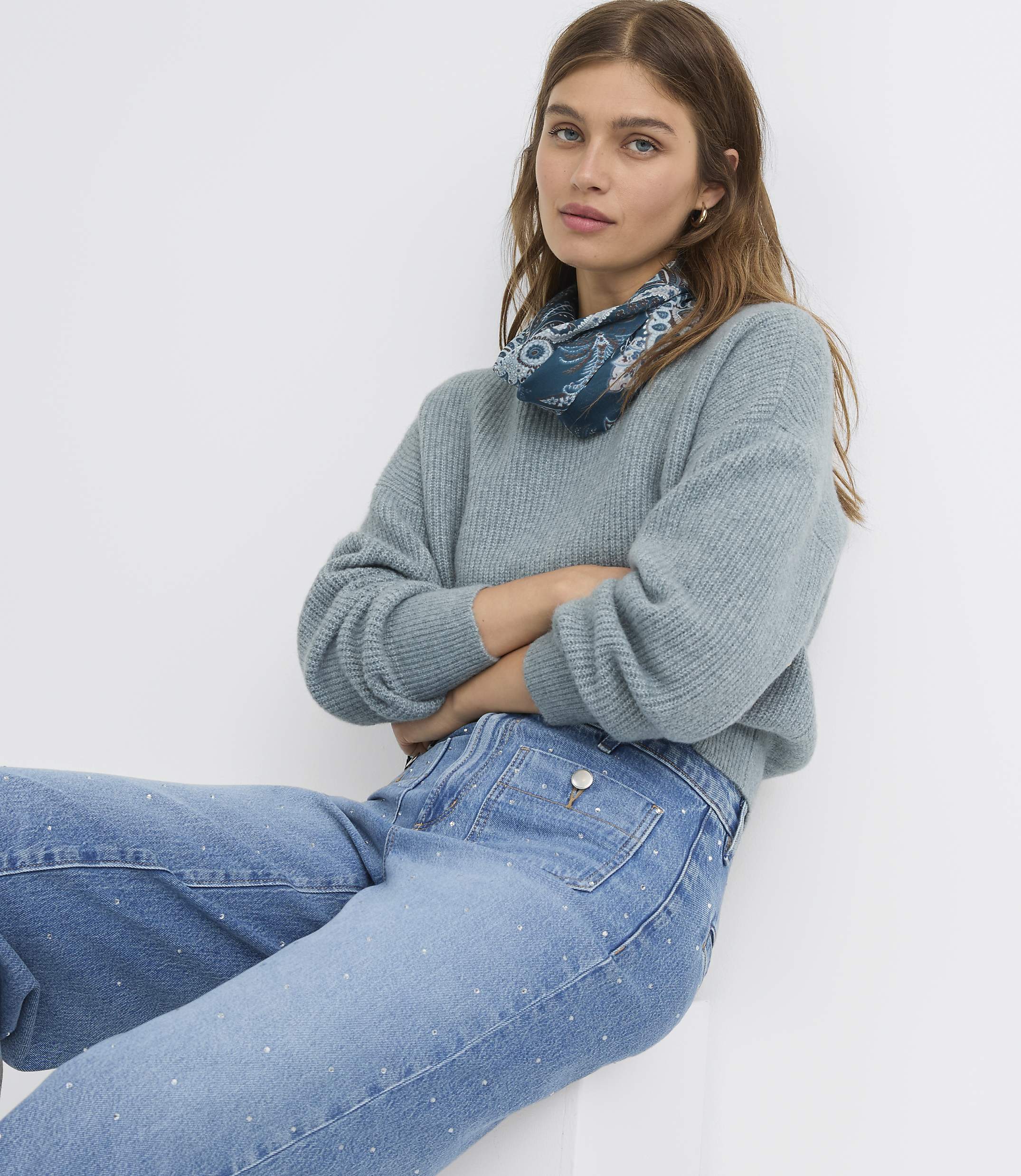 A woman in a light blue sweater and embellished jeans poses against a plain white background.