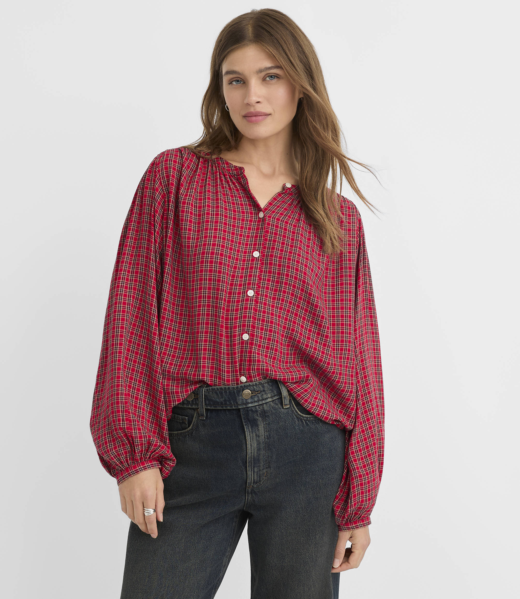 A woman in a red plaid blouse and dark jeans stands against a plain white background.