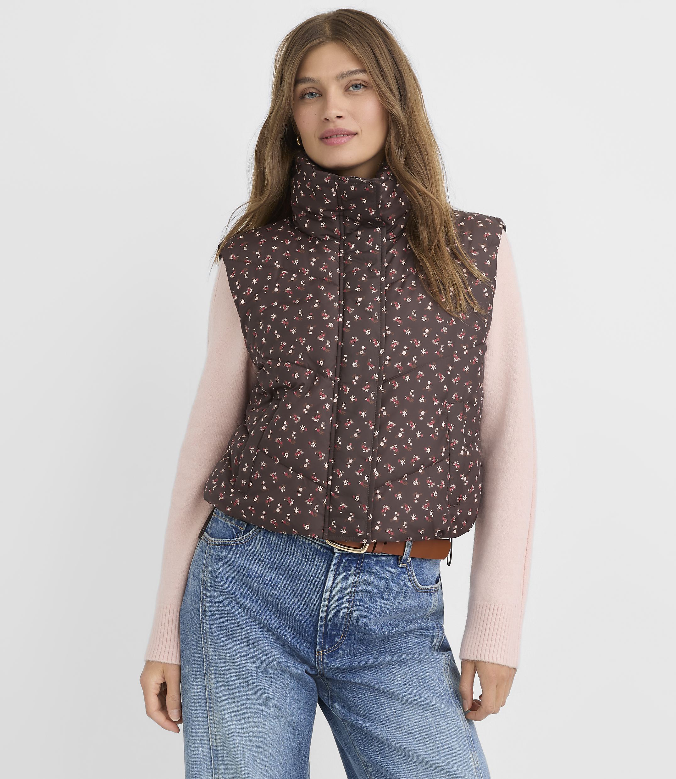 A woman in a floral puffer vest and pink sweater stands against a plain background.