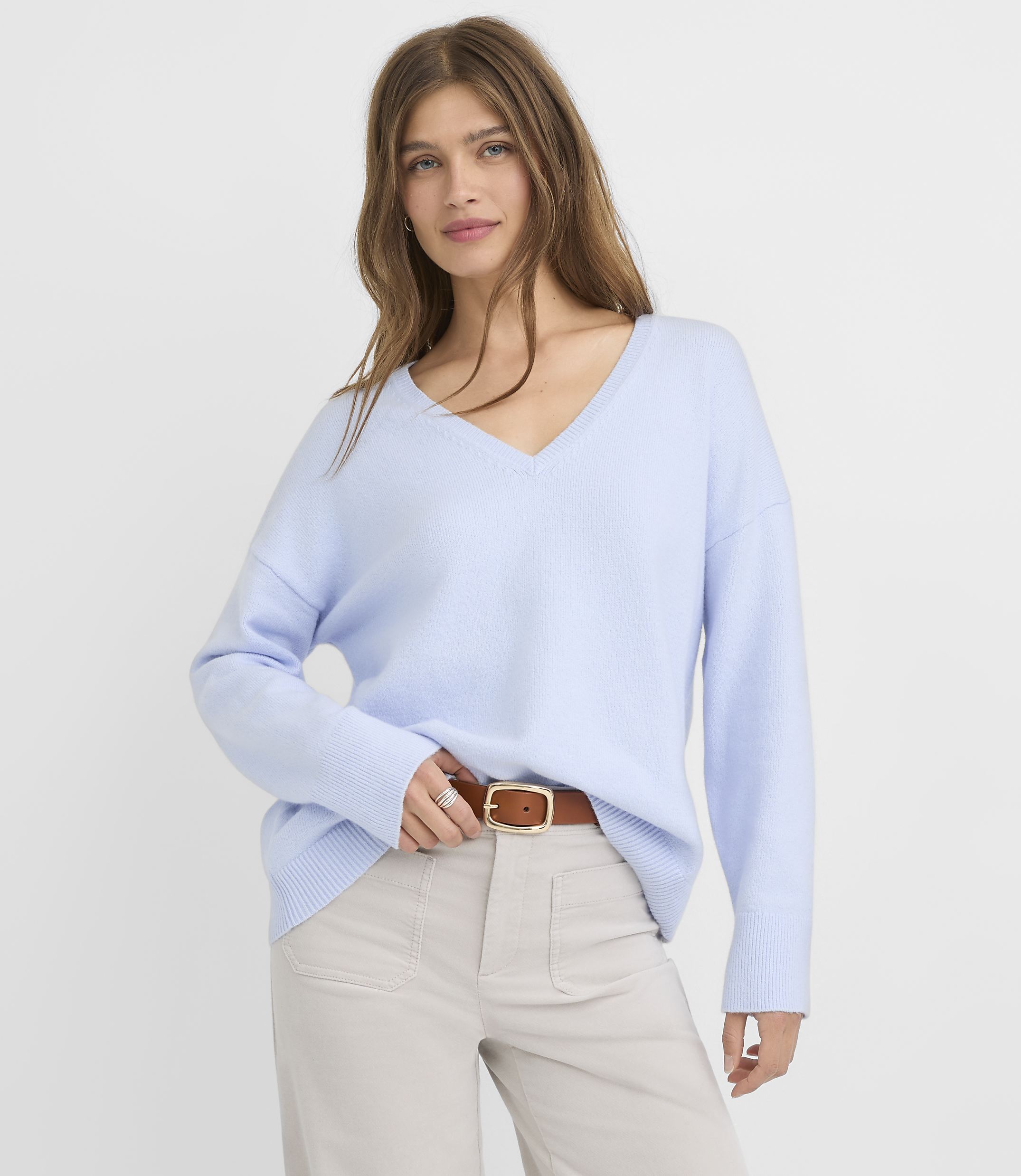 A woman in a light blue v-neck sweater and tan pants stands against a plain white background.