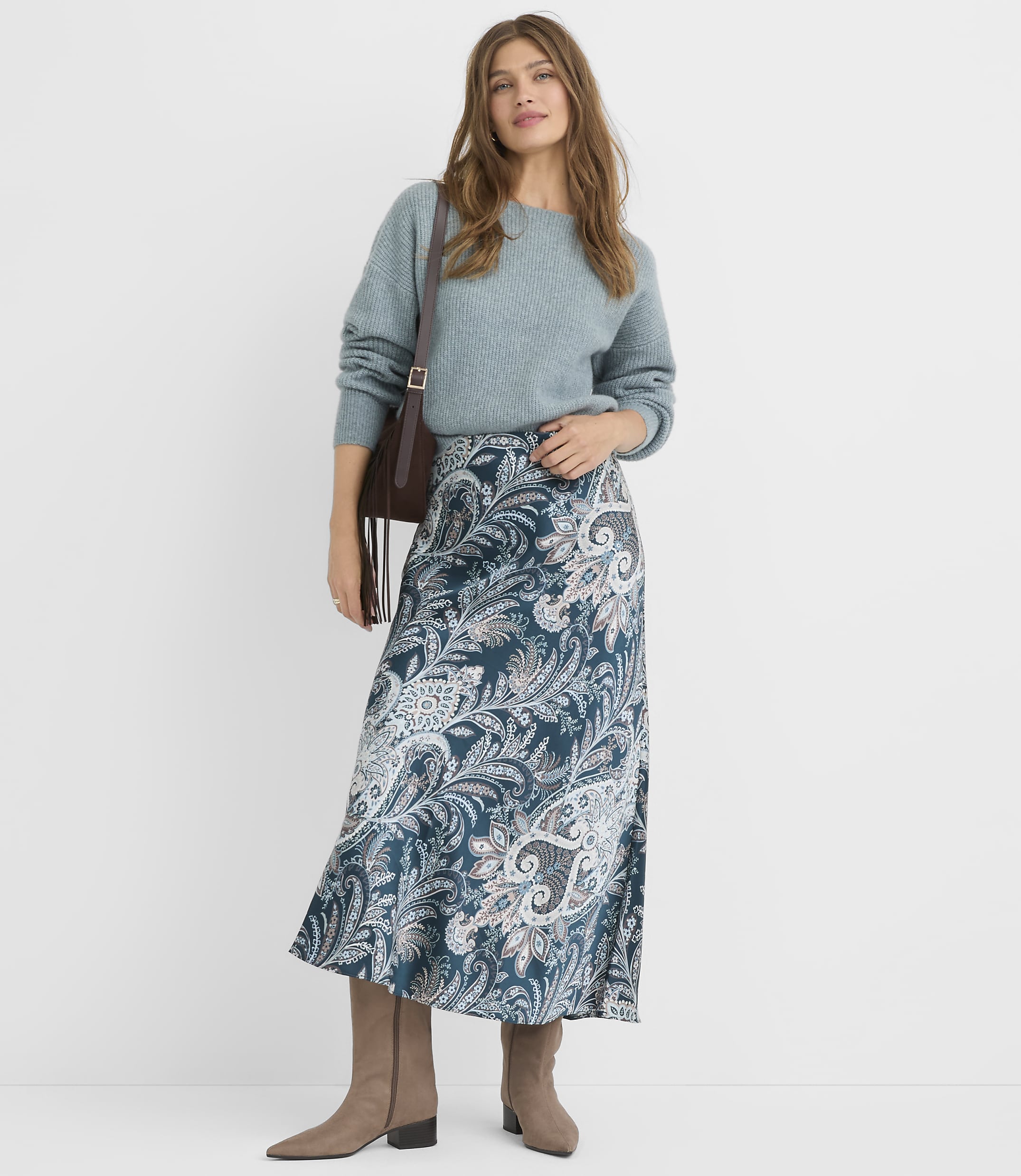 A woman in a teal sweater and a patterned skirt stands against a plain gray background.