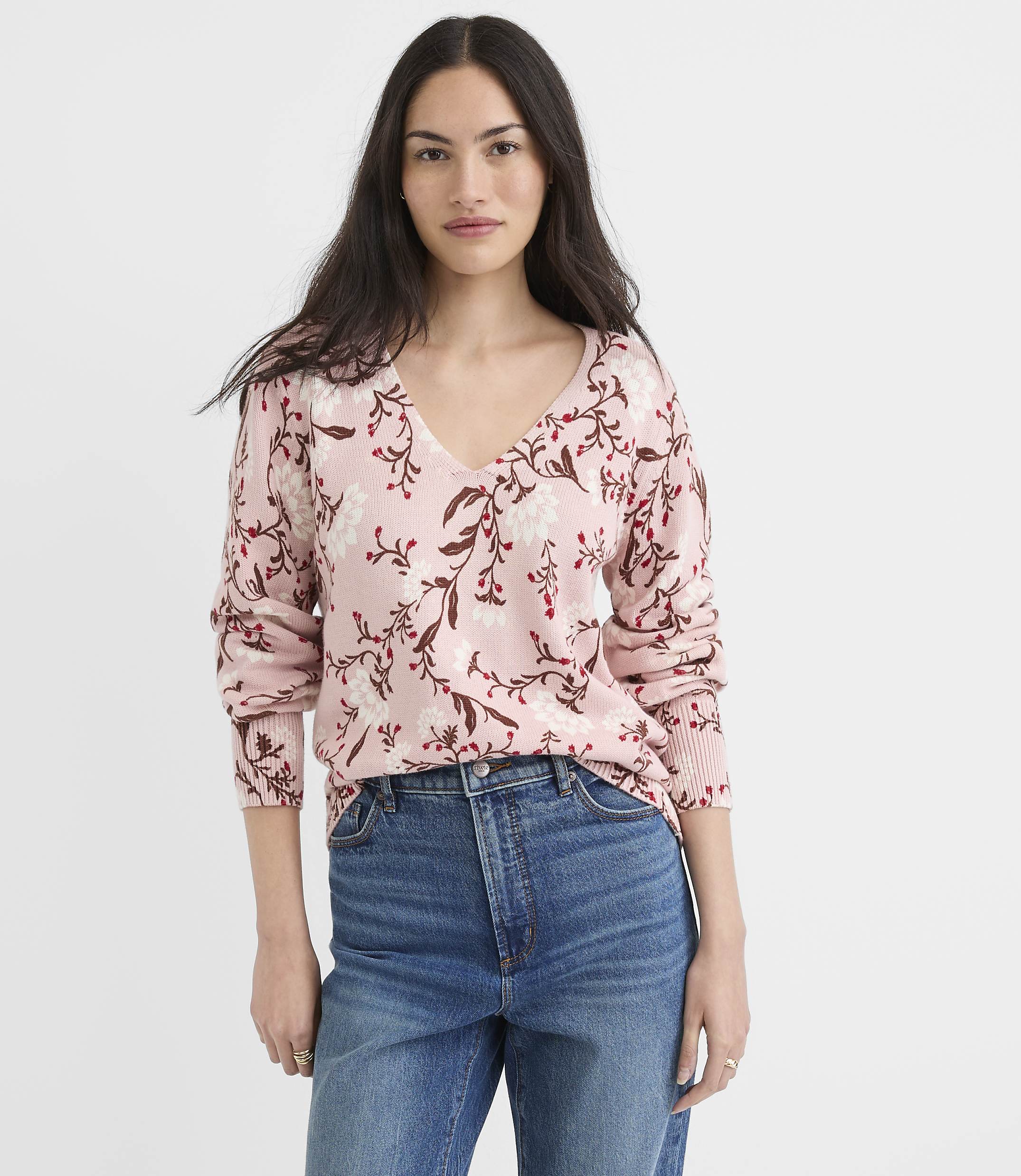 A woman in a floral pink sweater and blue jeans stands against a plain white background.