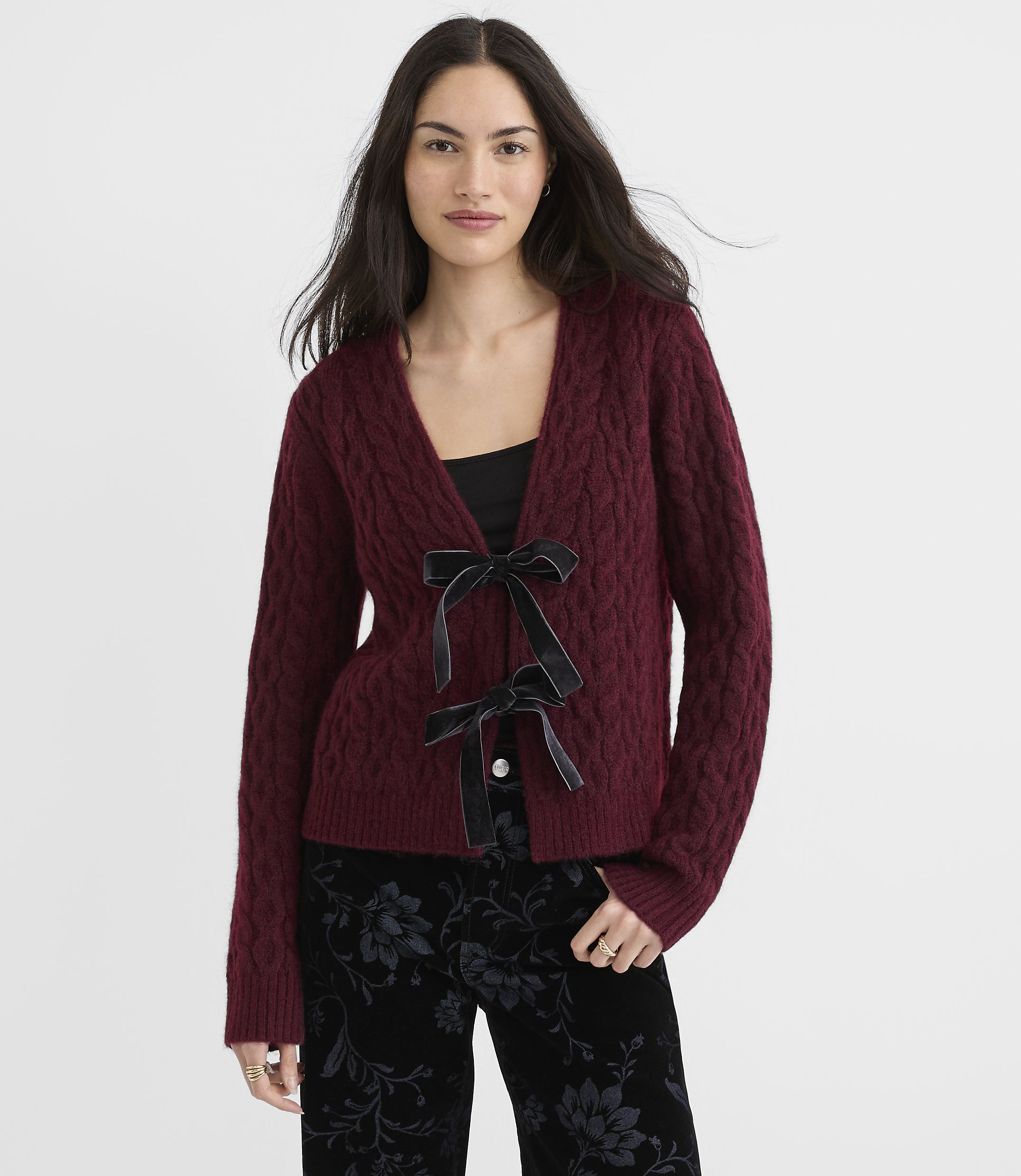 A woman in a burgundy cable-knit cardigan with black velvet bows stands against a dark, patterned background.