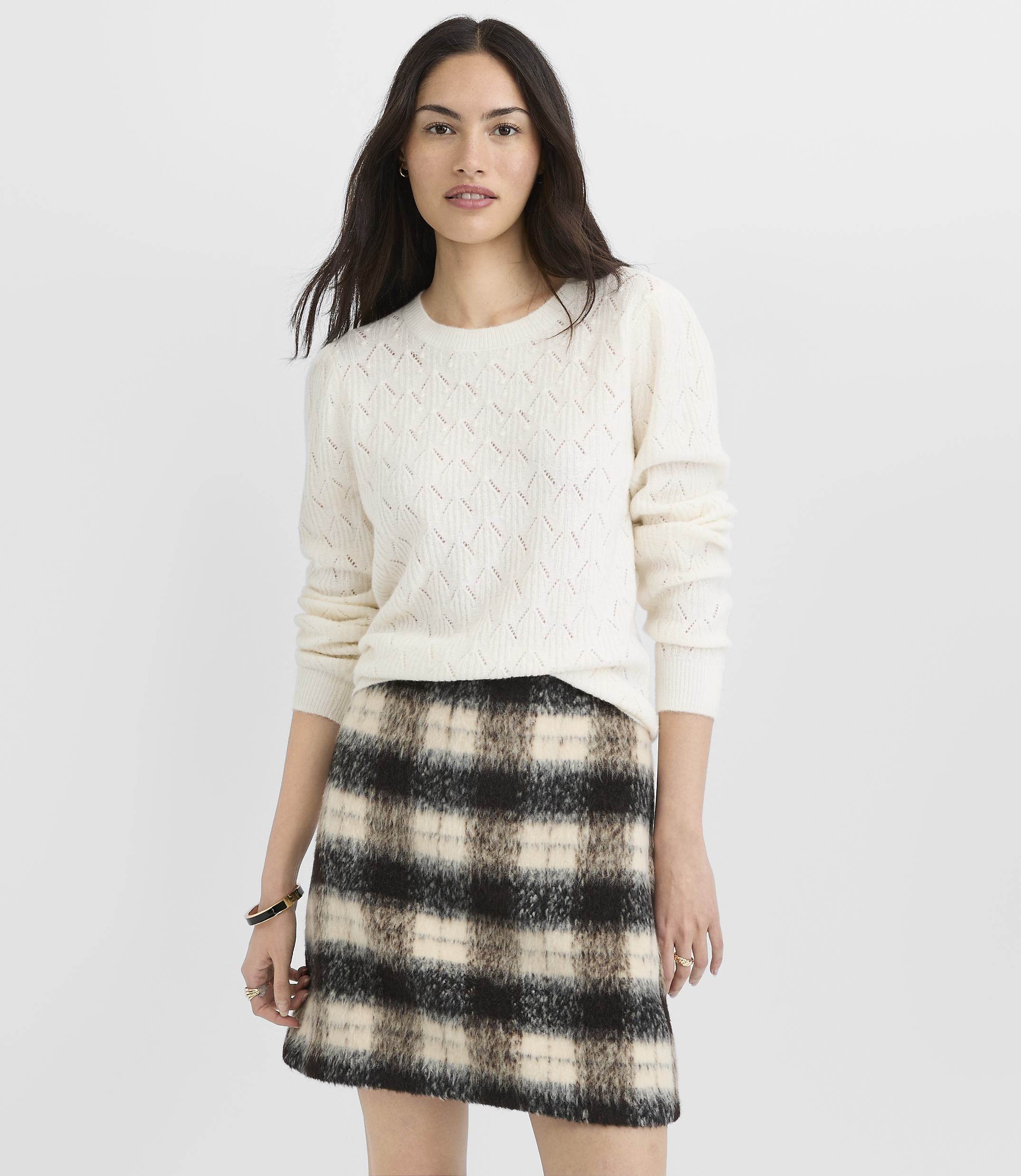 A woman in a cream cable-knit sweater and a plaid mini skirt stands against a plain white background.