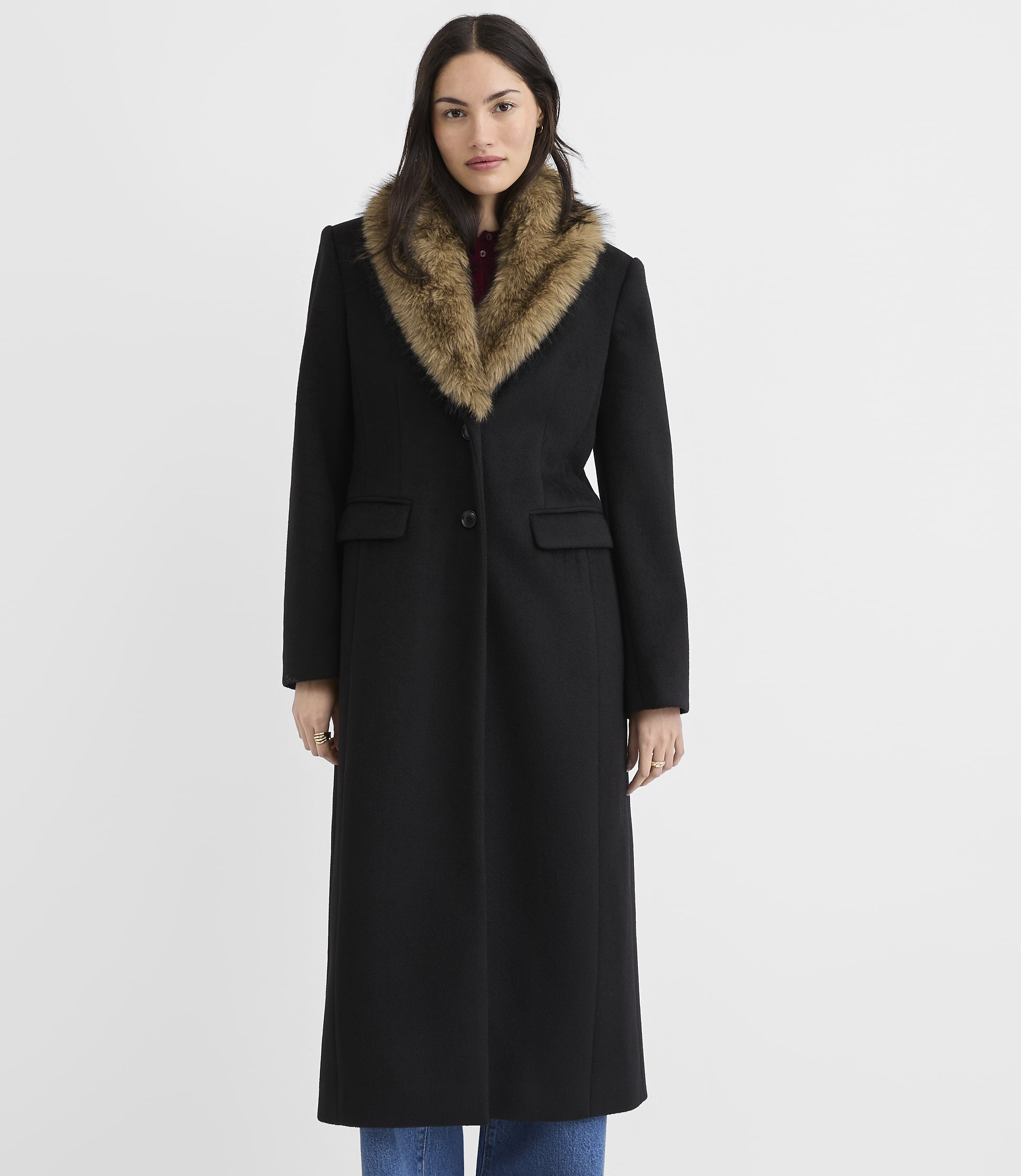 A woman in a long black coat with a fur collar stands against a plain white background.