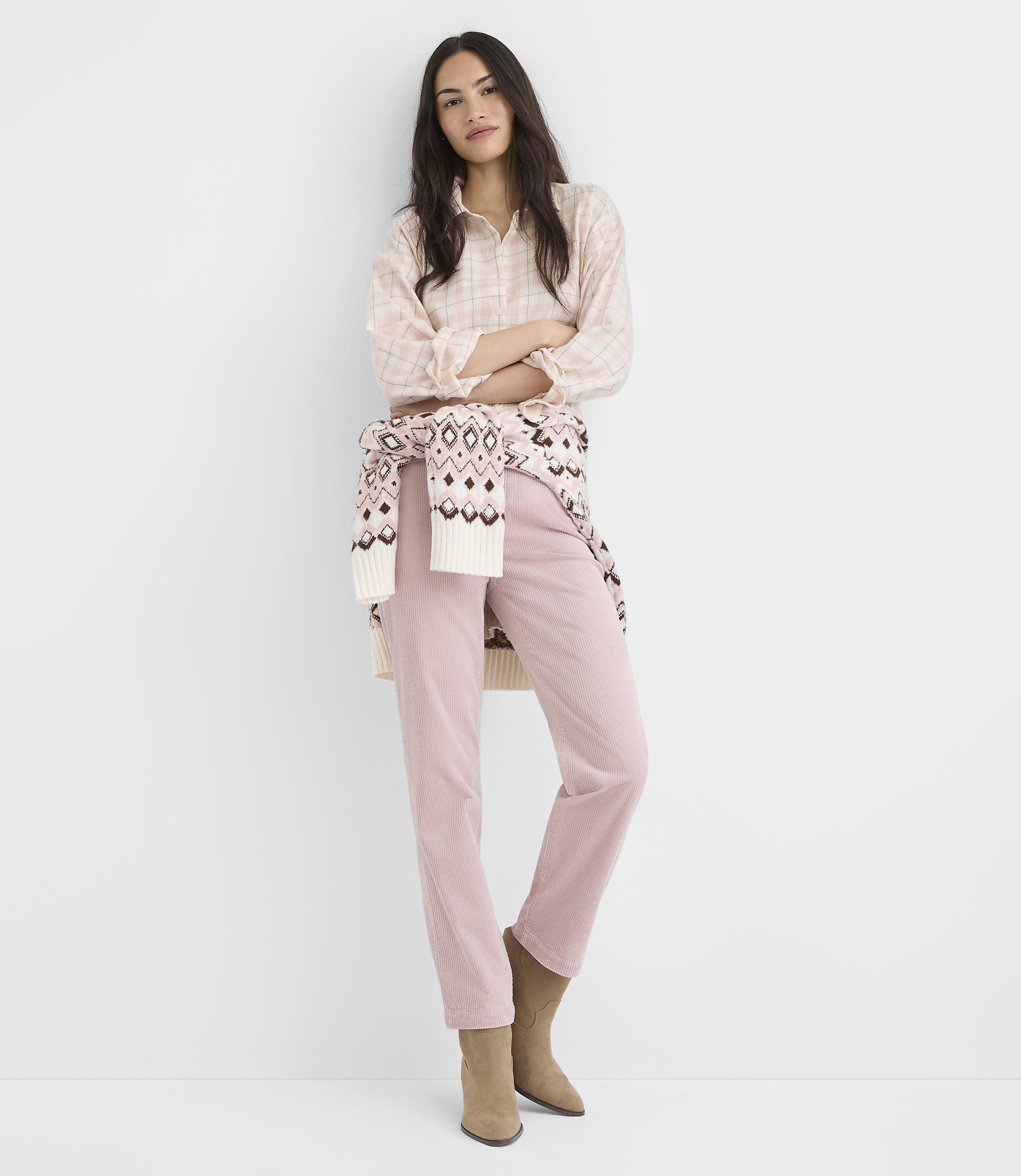 A woman in a patterned shirt, pink corduroy pants, and boots stands with her arms crossed against a plain white background.
