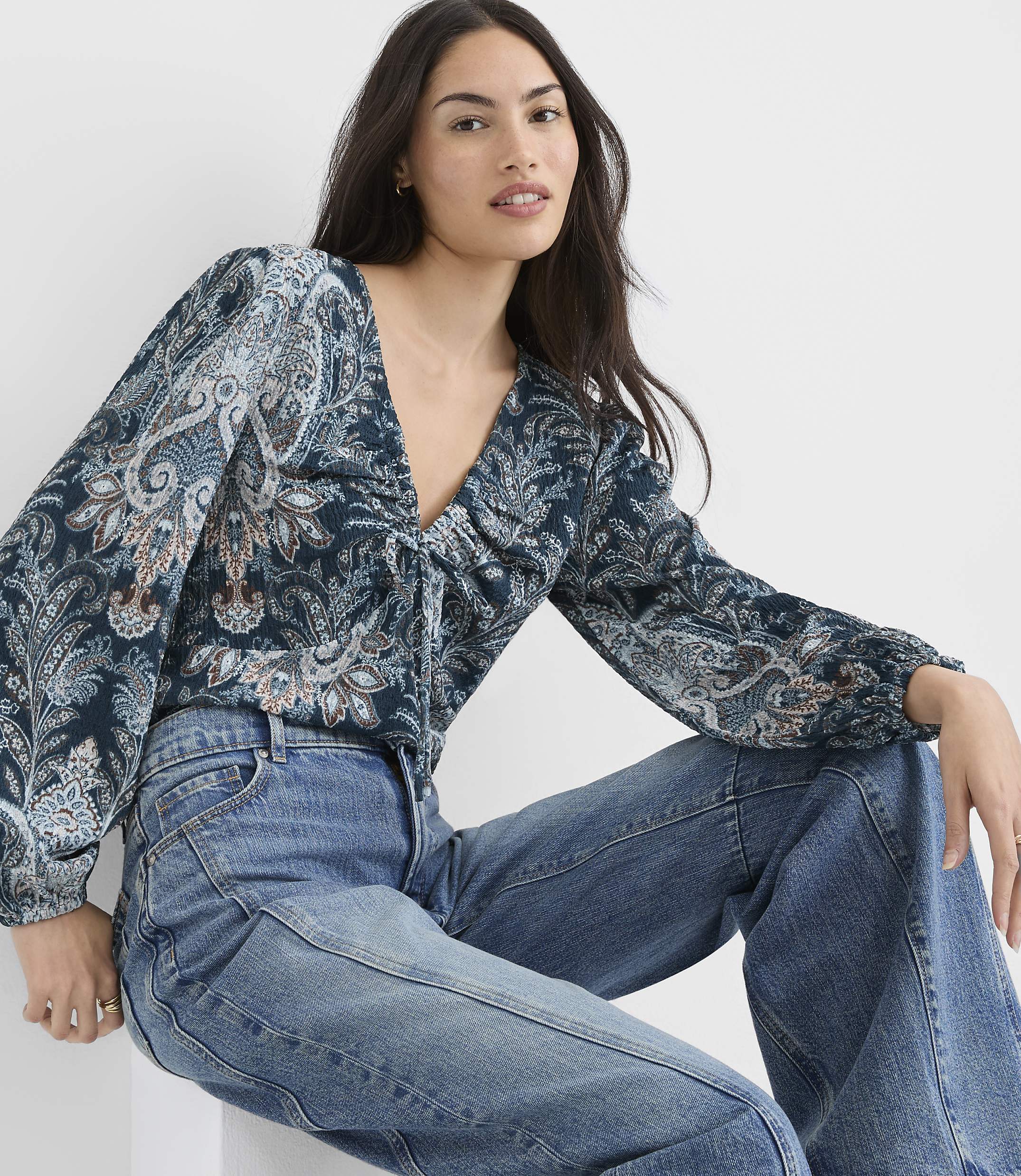 A woman in a patterned blouse and jeans sits against a plain white background.